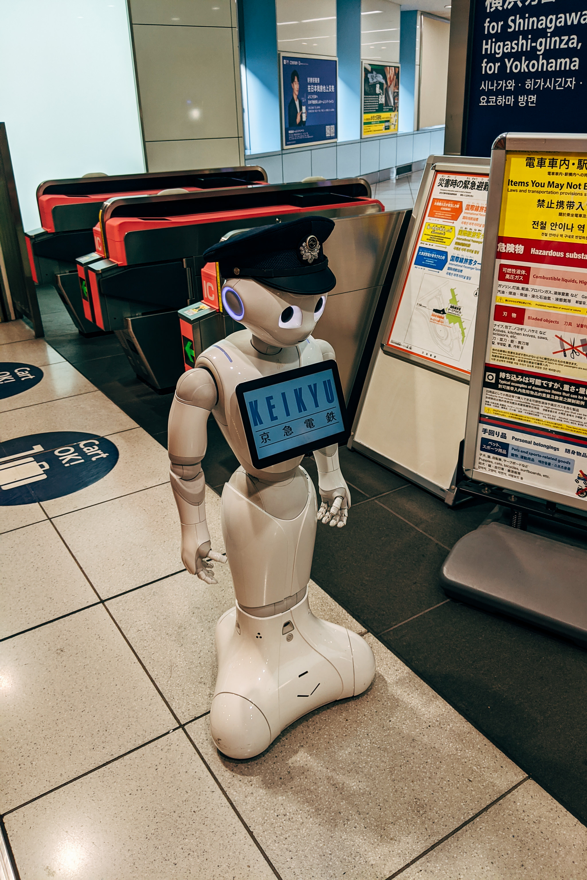 Robot in a train station wearing a police hat, holding a sign that says 'KEIKYU' in English and Japanese.
