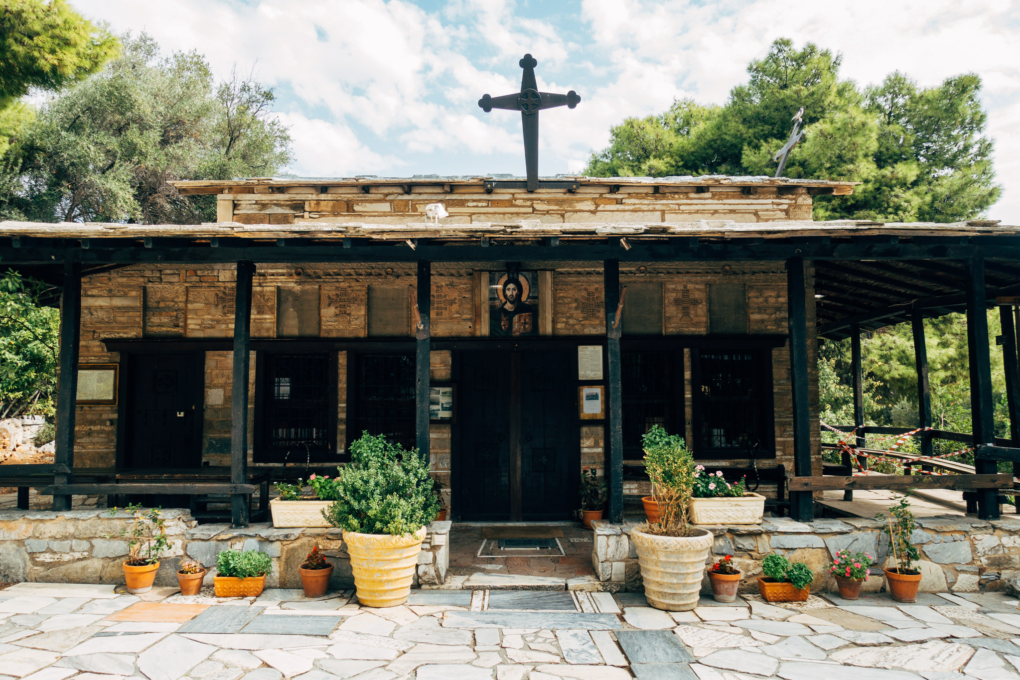 Stone church with a dark wooden portico, a large cross on the roof, and potted plants.