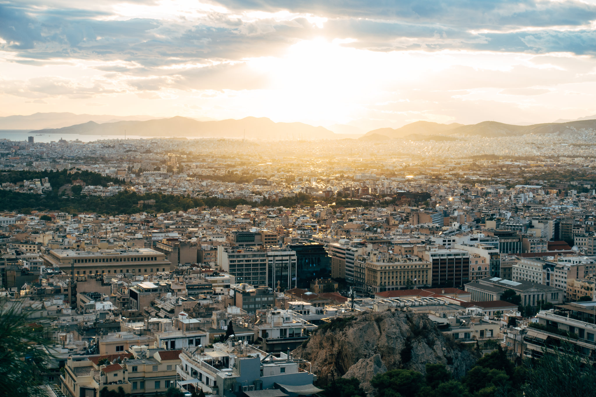 Sunset view of Athens, Greece from a hilltop, showing the city sprawling out towards the sea and mountains in the distance.