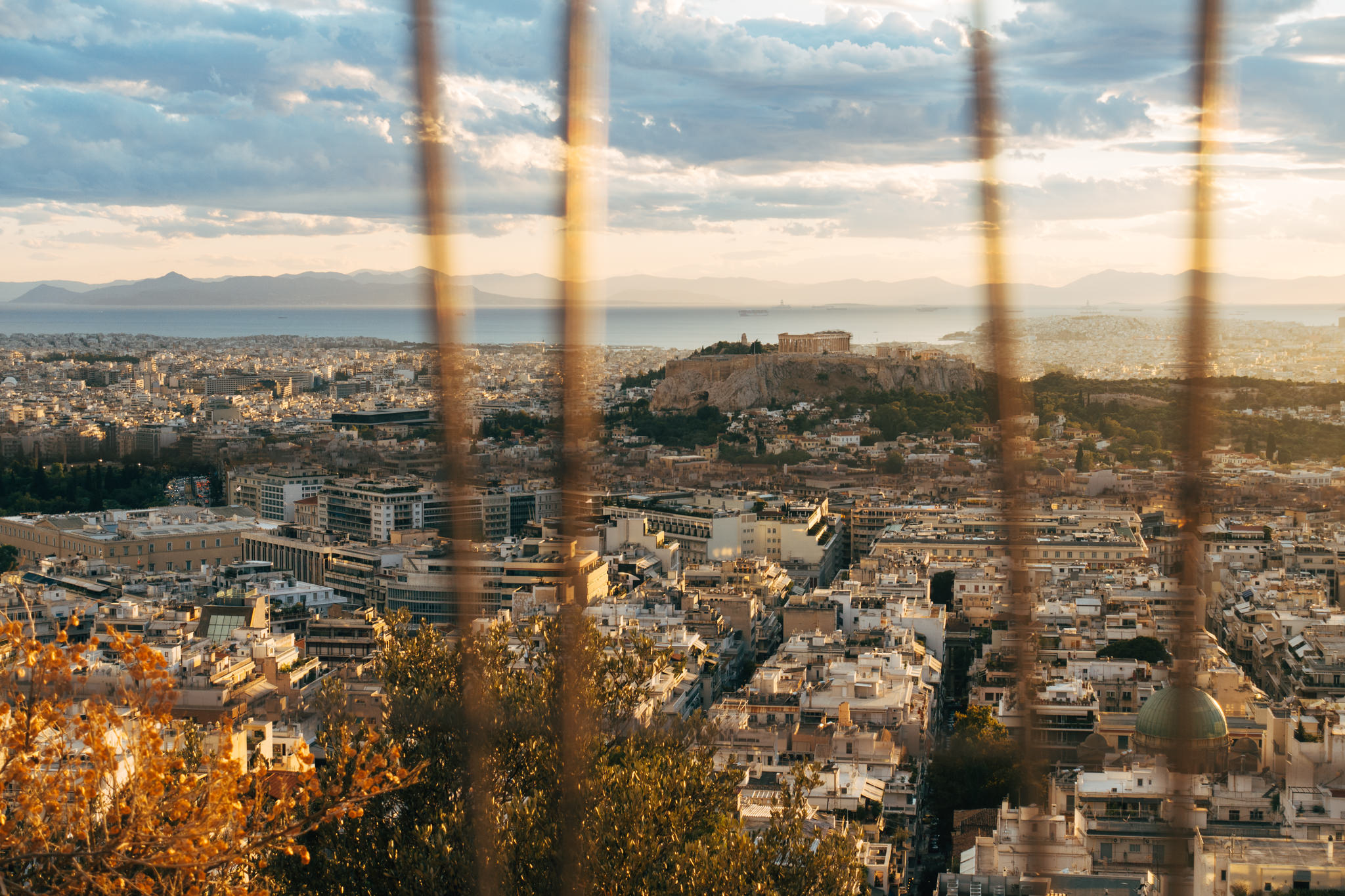 Athens cityscape with Acropolis and Mount Lycabettus in the background, viewed through a metal fence.