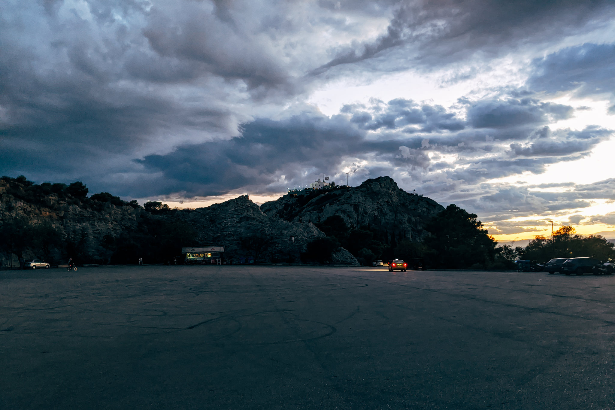 Mount Lycabettus at dusk, viewed from an empty parking lot with a few cars and a small building.