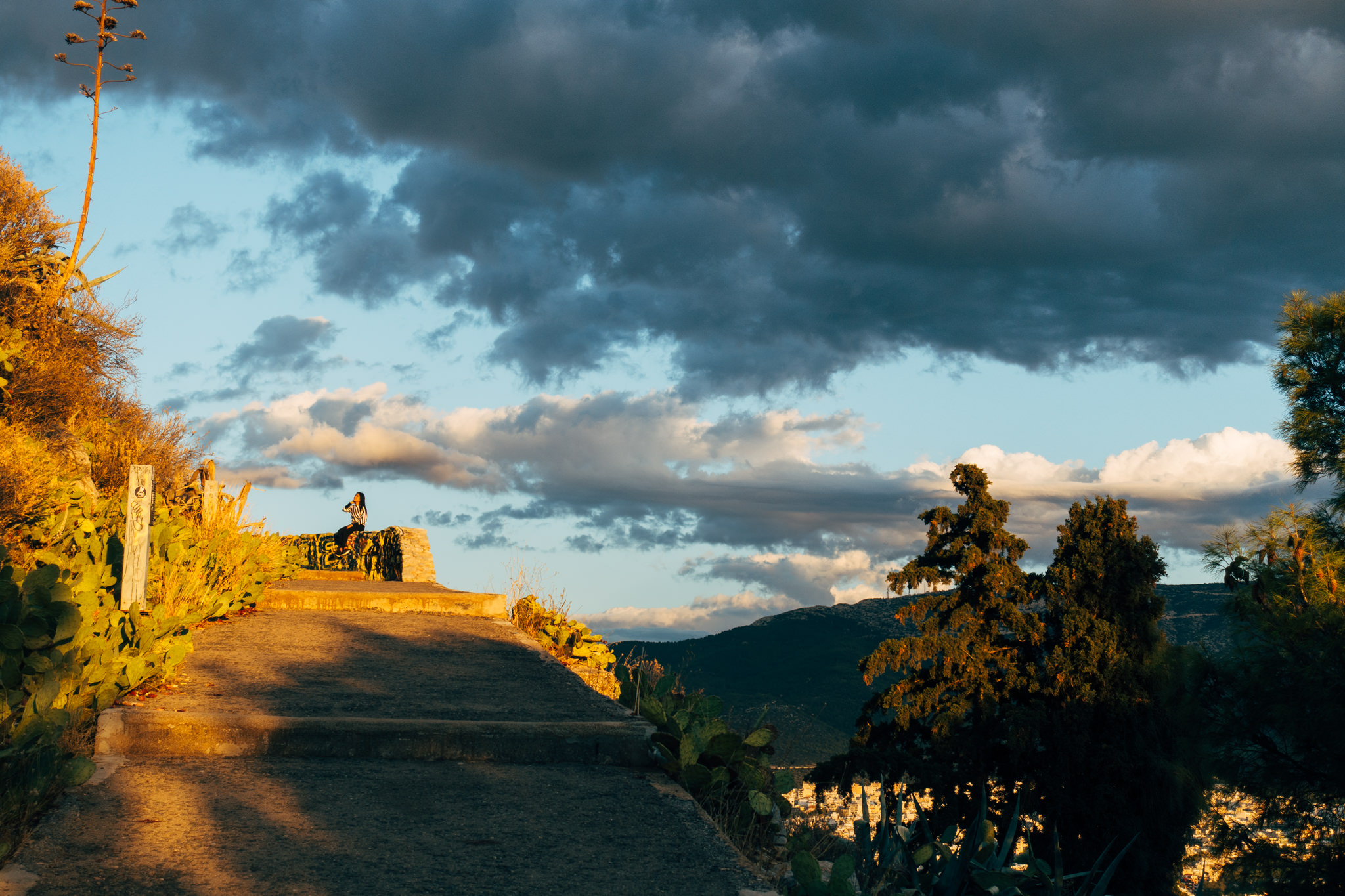Person sitting on a stone wall overlooking a city under a dramatic sky.