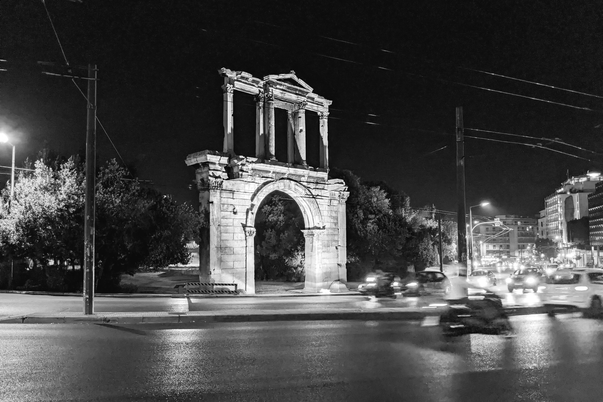 Black and white photo of the Arch of Hadrian in Athens at night, with blurred traffic in the foreground.