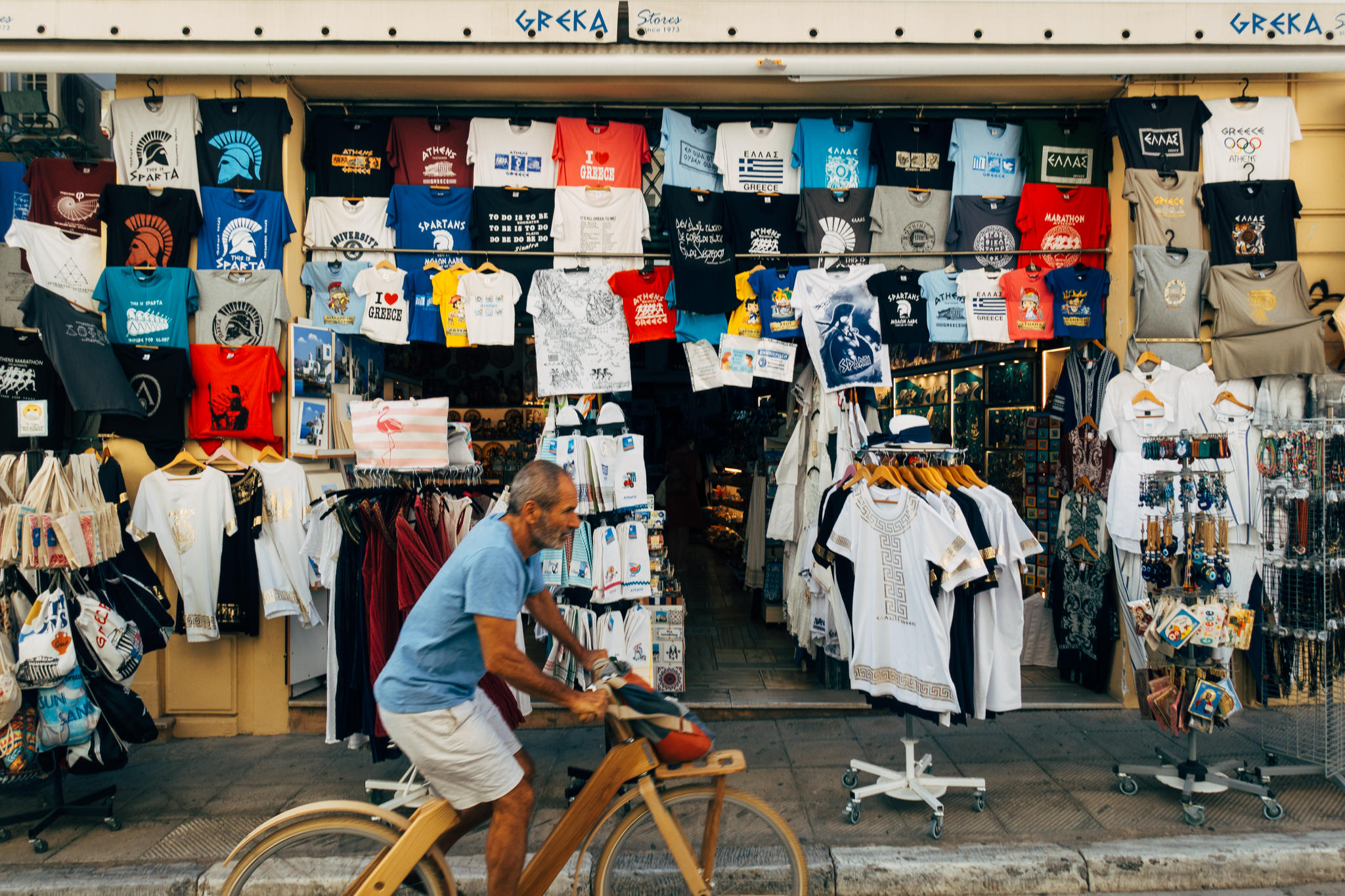 Man on bicycle passing tourist shop in Athens, Greece, displaying Greek-themed t-shirts and souvenirs.