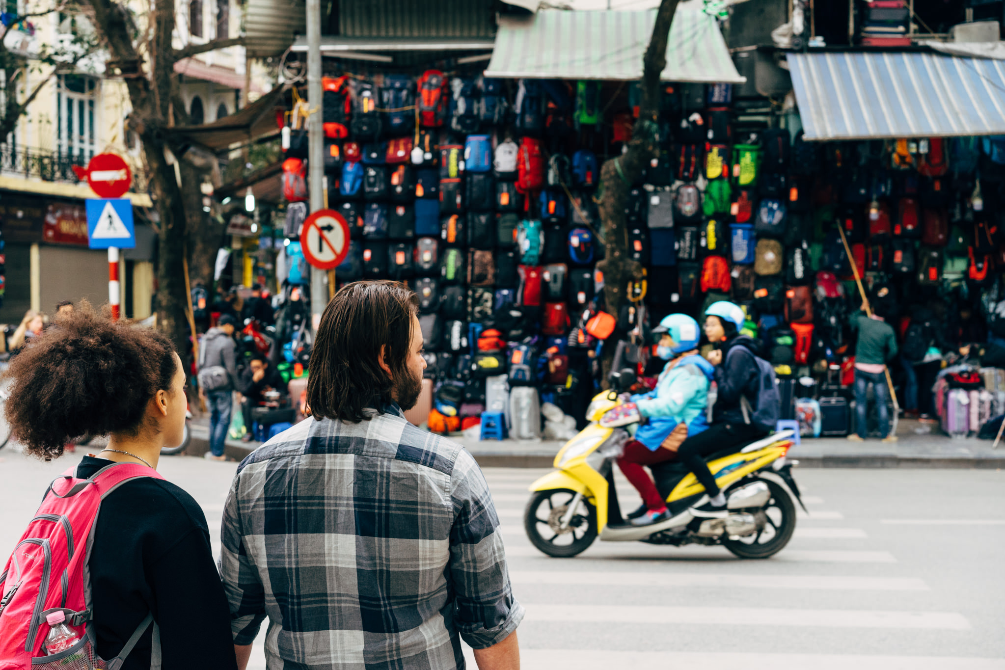 Couple crossing street in Hanoi, Vietnam, with a scooter and a backpack shop in the background.