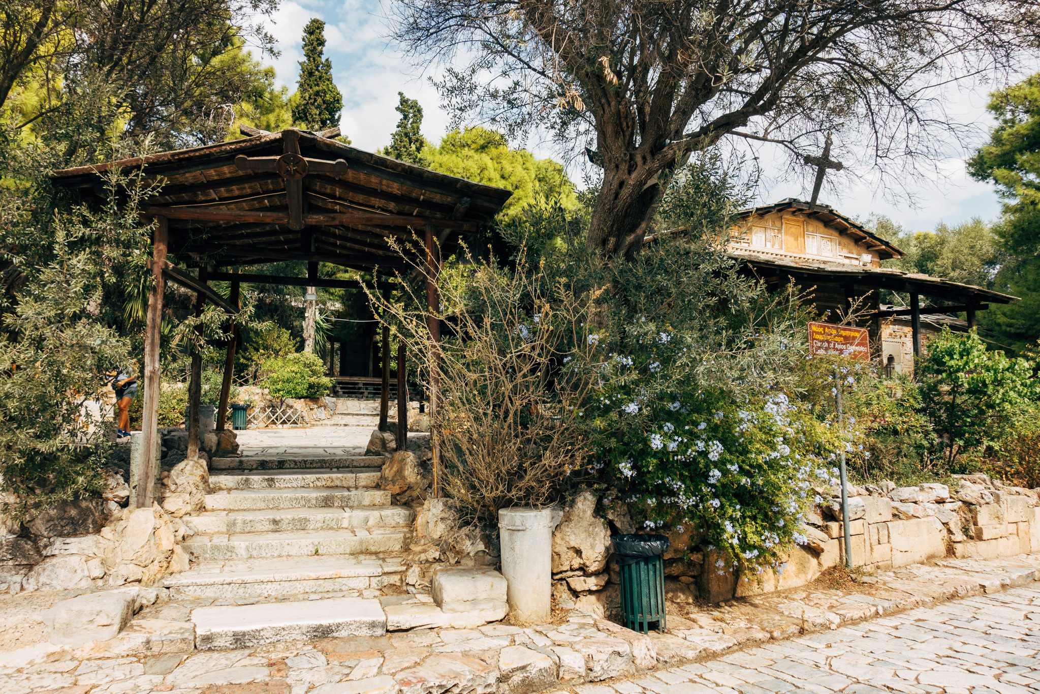 Stone steps leading to a wooden pergola and the Church of Ayios Demetrios Loumbardiaris in a garden setting.
