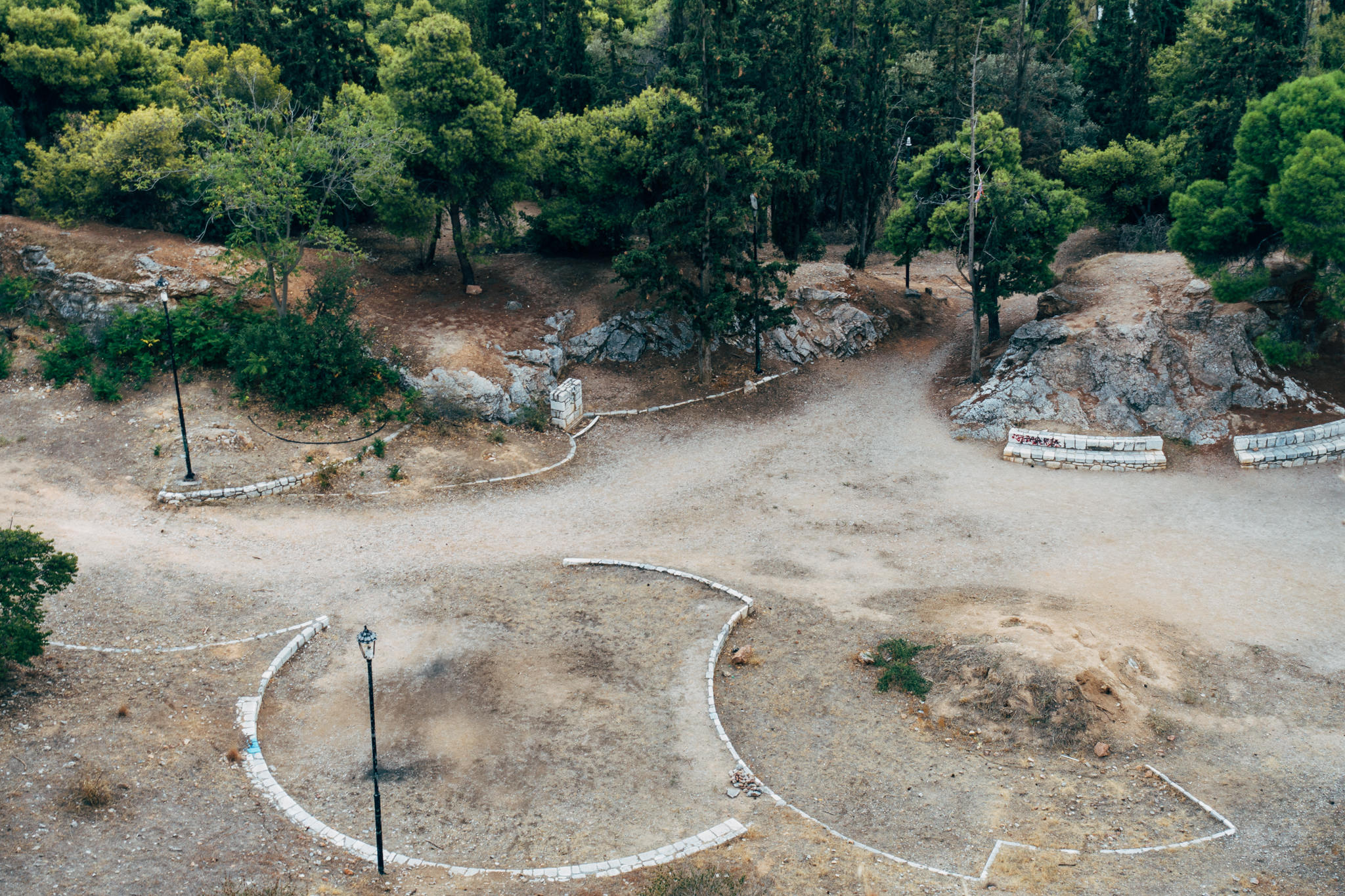 Aerial view of Filopappou Hill in Athens, showing a dry, rocky landscape with stone benches and lampposts.