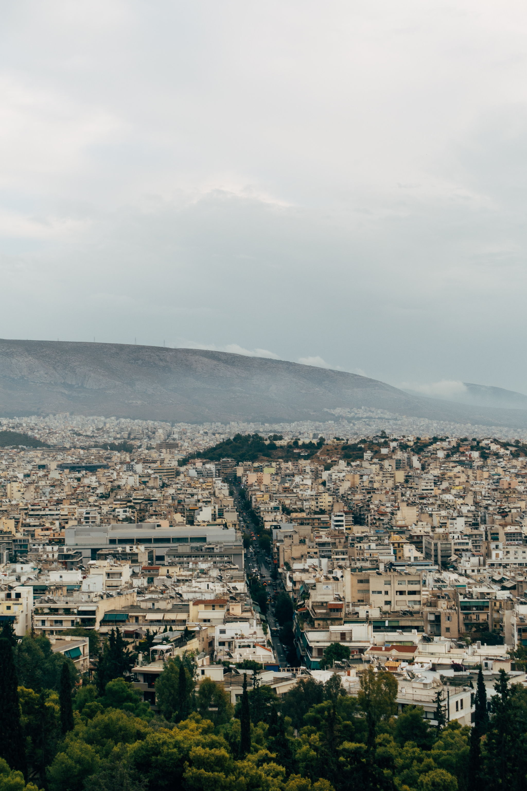 Aerial view of Athens, Greece, showing a dense cityscape with a street running through it, and a mountain in the background.