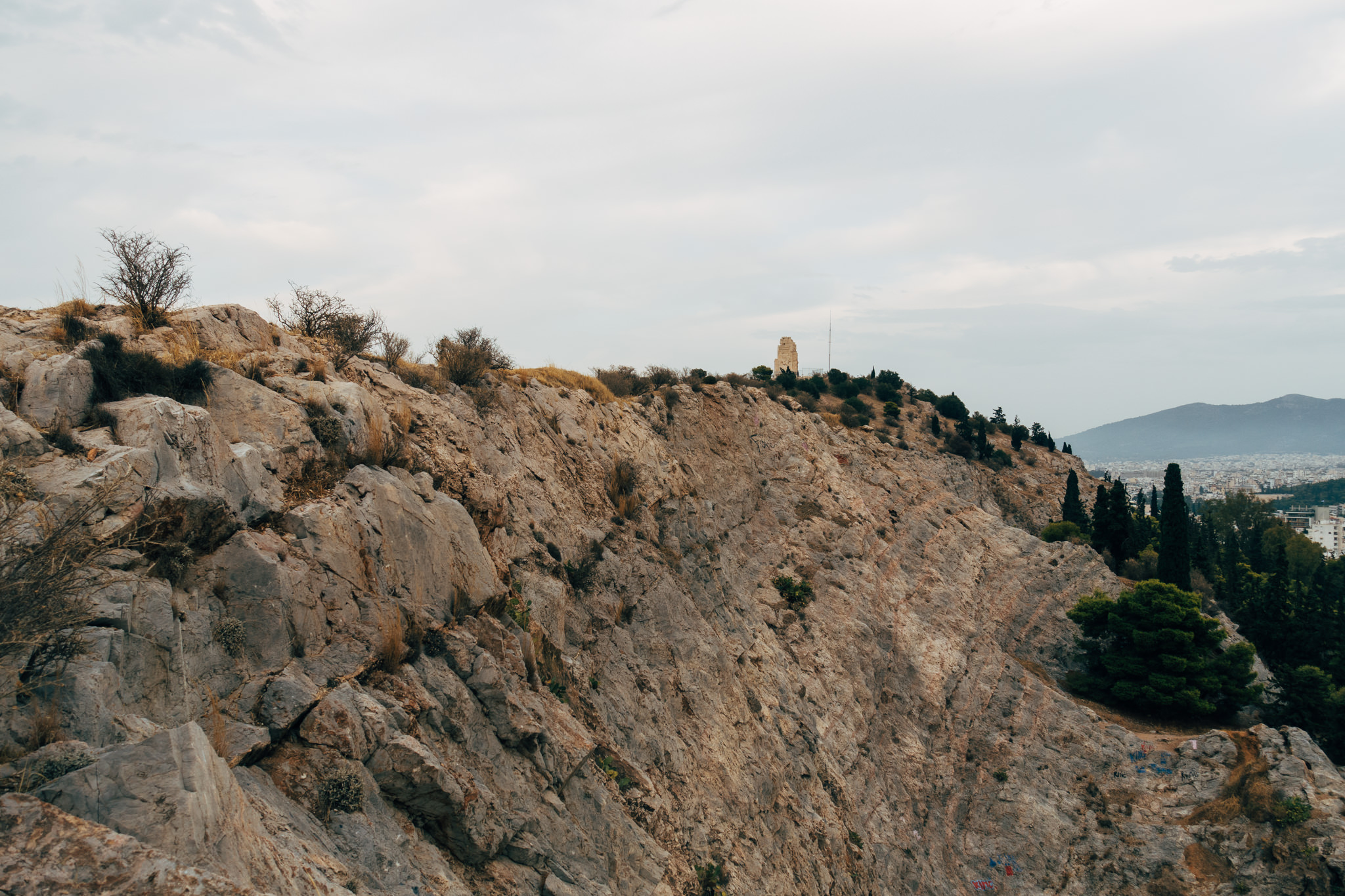 Filopappou Hill in Athens, showing a rocky cliff face with sparse vegetation and a monument visible in the distance.