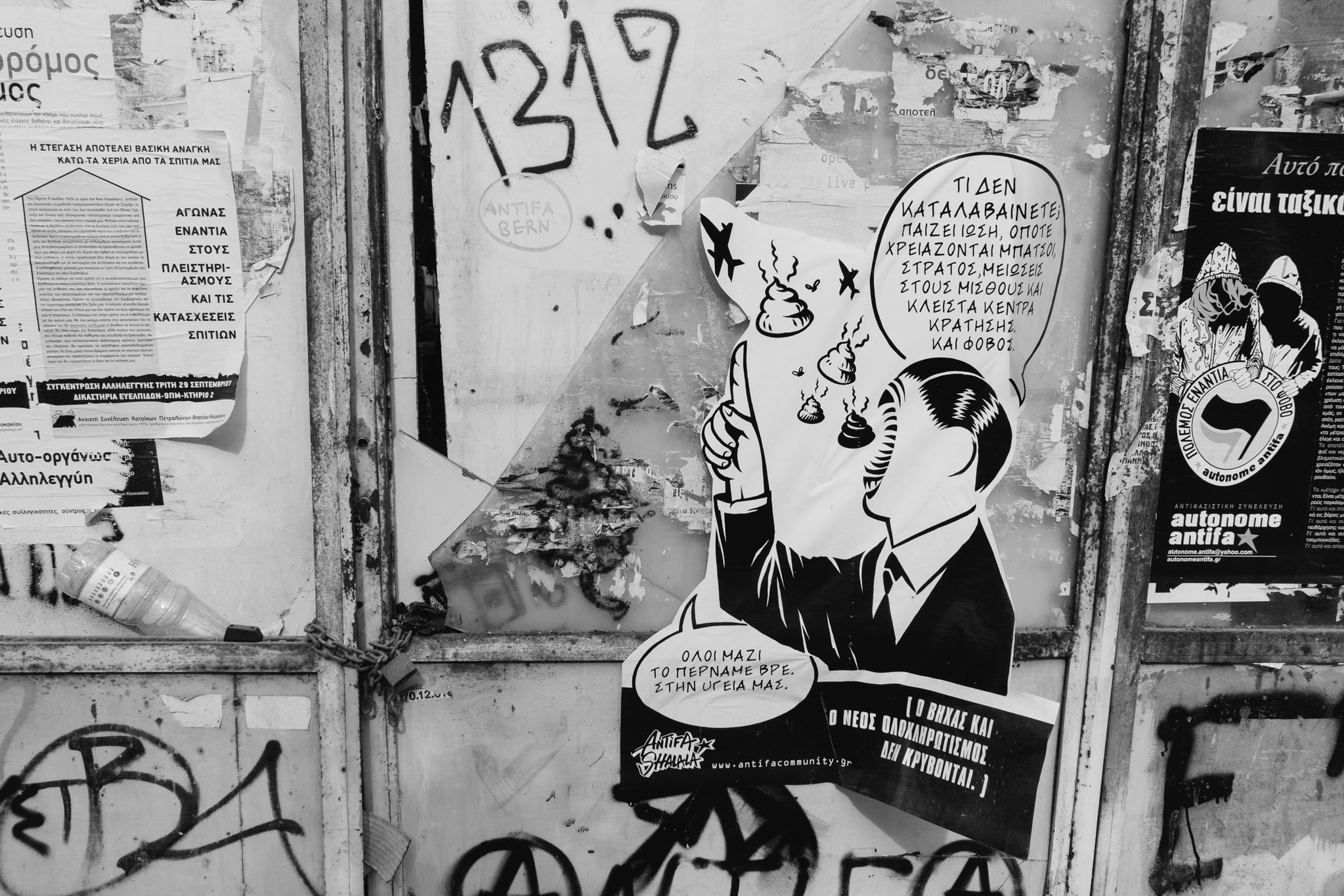 Black and white photo of a wall covered in graffiti and posters in Athens, Greece, featuring a sticker depicting a man being offered feces.