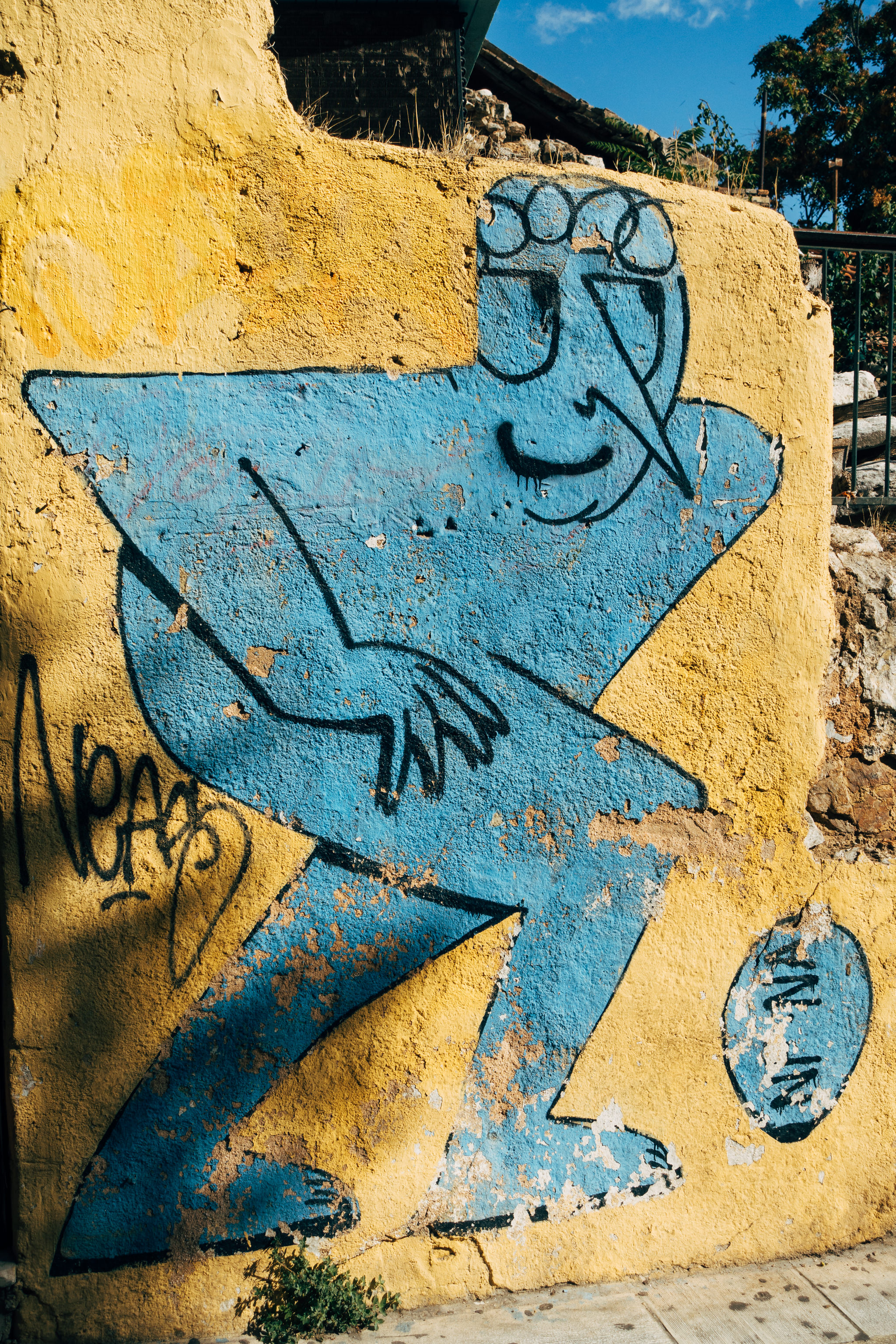 Graffiti of a blue figure playing soccer on a weathered yellow wall in Athens, Greece.