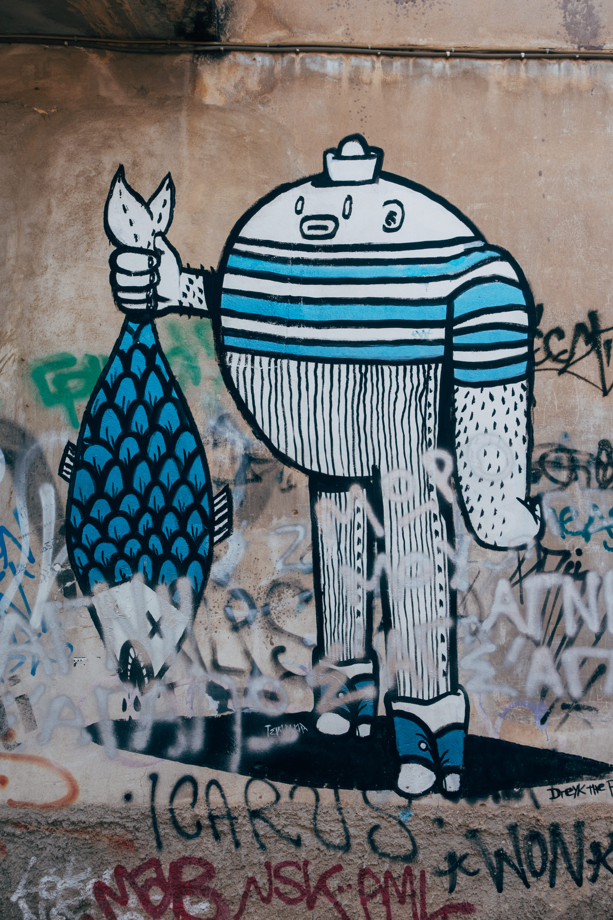 Graffiti of a cartoon figure holding a large blue fish on a textured wall with other graffiti.
