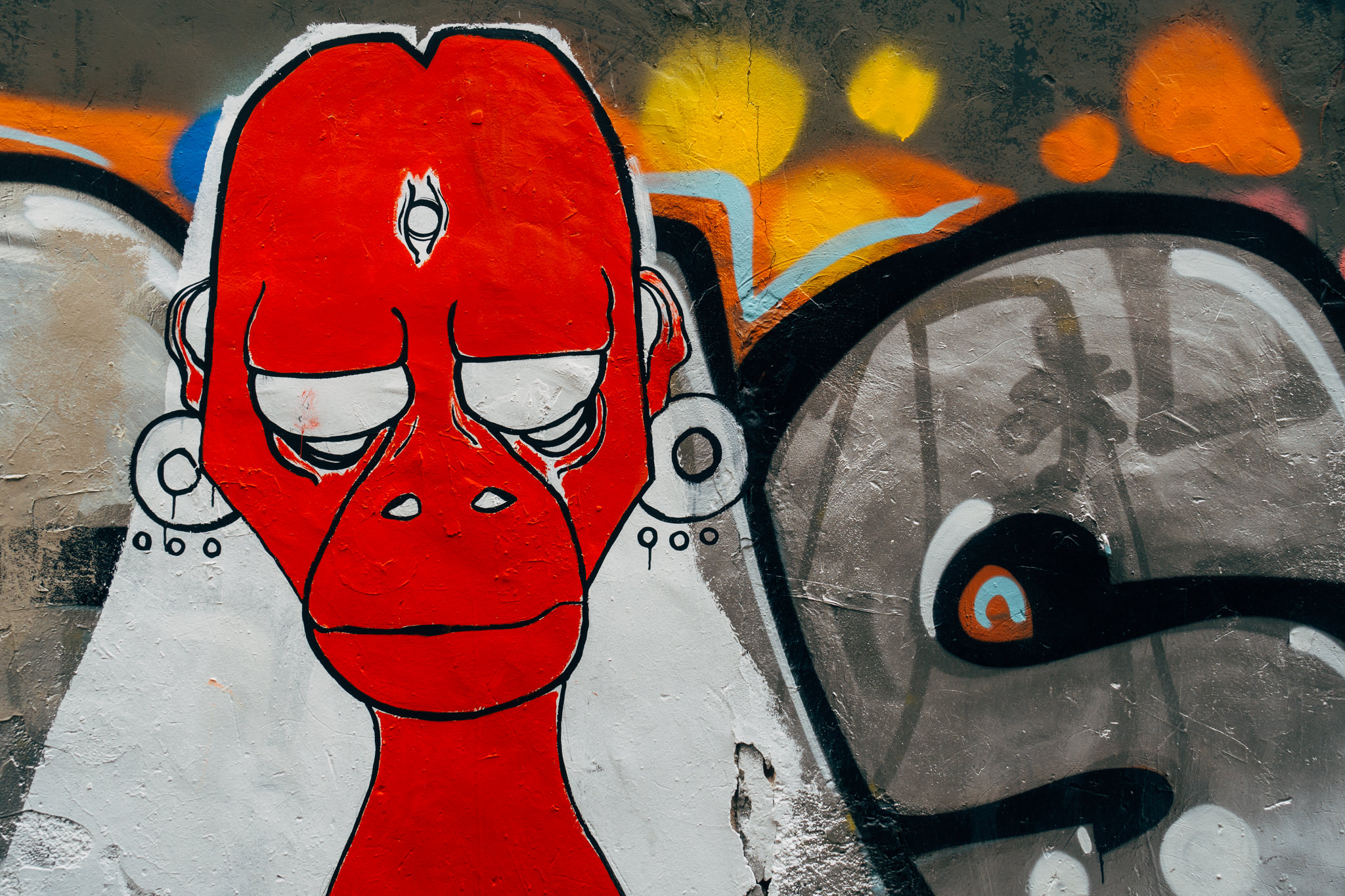 Graffiti of a red face with a third eye, large earrings, and closed eyes on a wall with other graffiti.