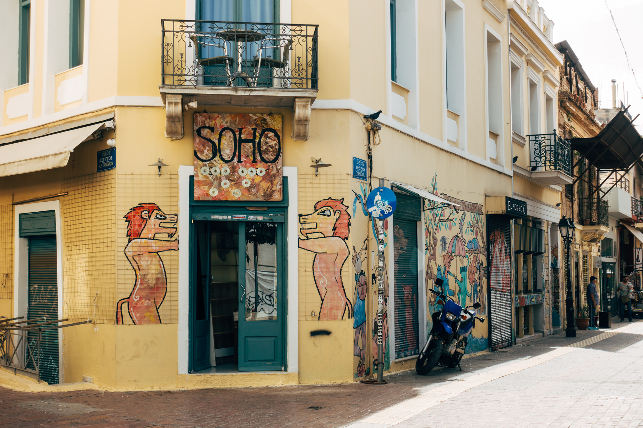 Graffiti-covered building corner in Athens, Greece, with a Soho shop and a motorcycle parked outside.