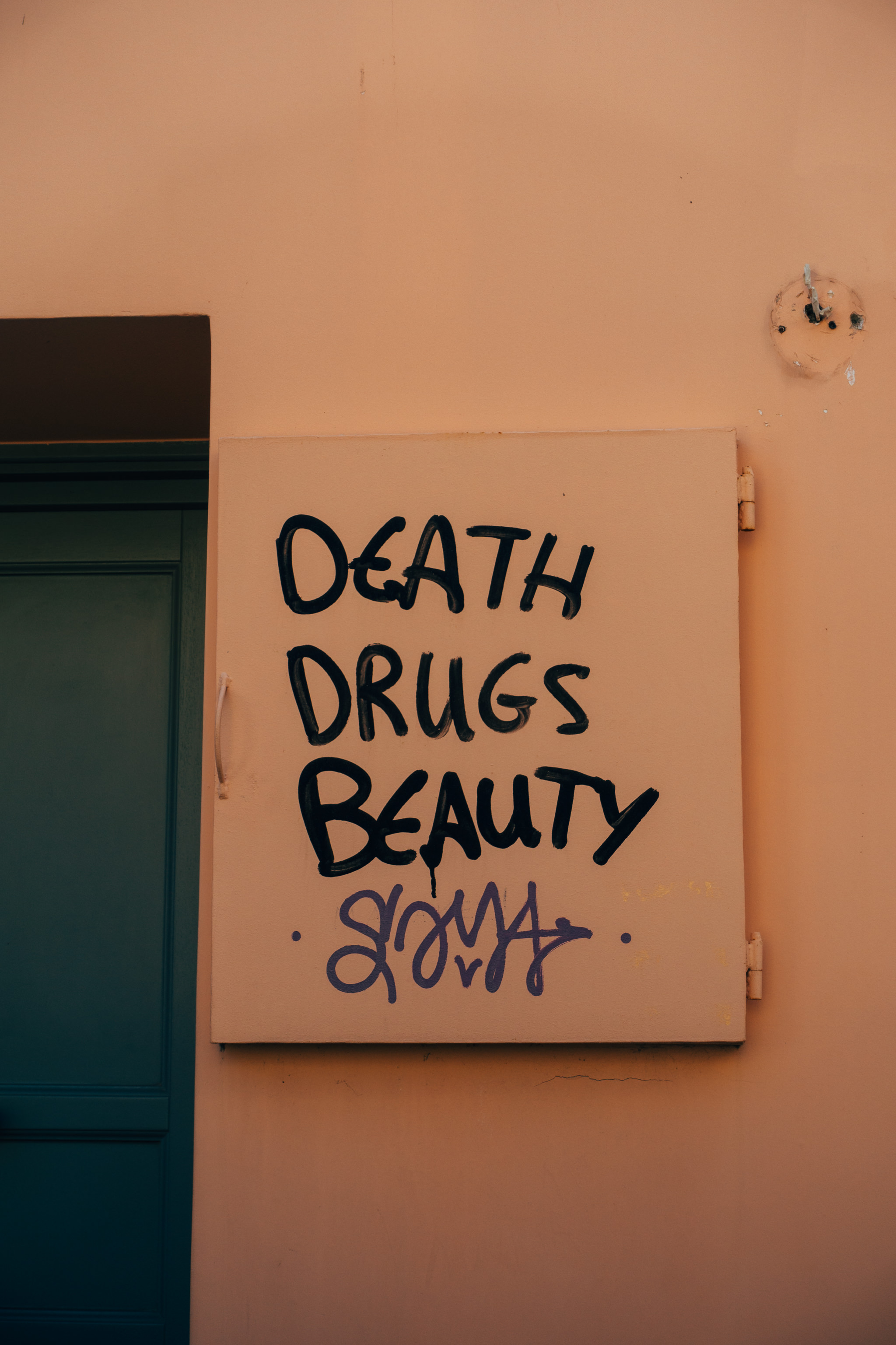 Graffiti on a tan wall reads 'DEATH DRUGS BEAUTY' in black, with smaller purple graffiti below.