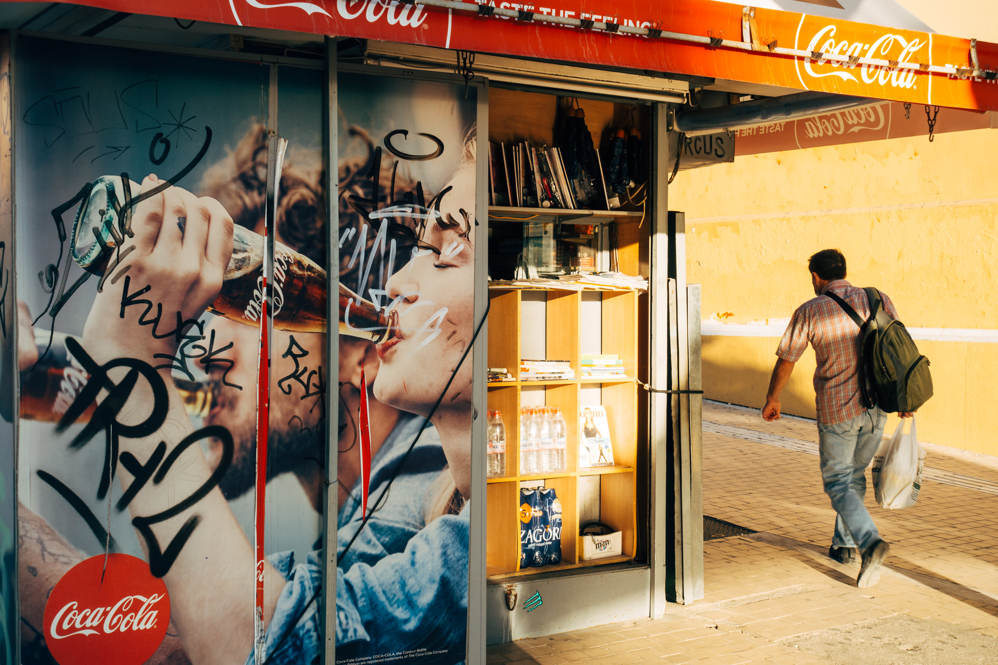 Graffiti-covered Coca-Cola advertisement on a kiosk; man walks away.