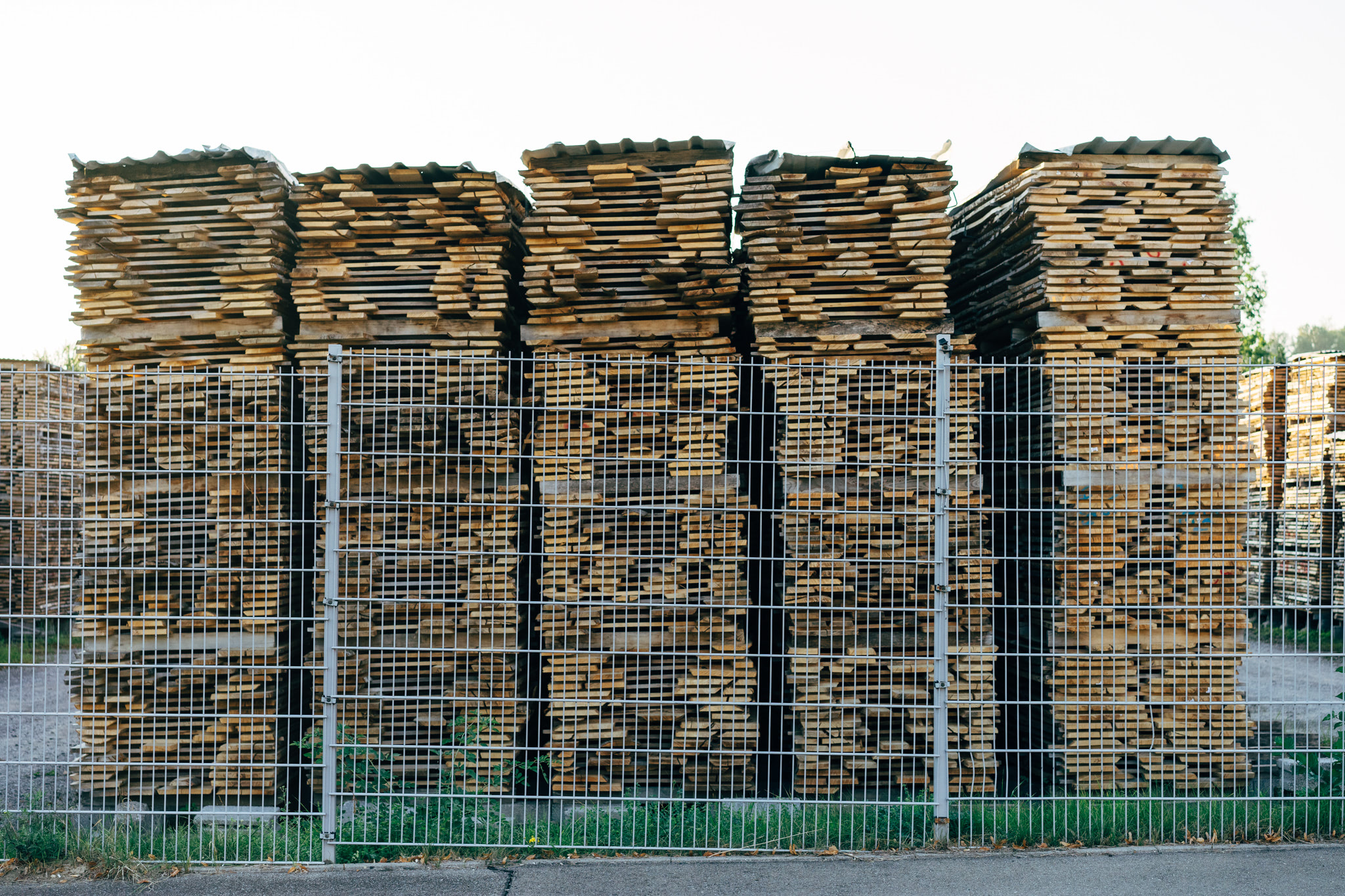 Four large stacks of lumber behind a chain link fence.