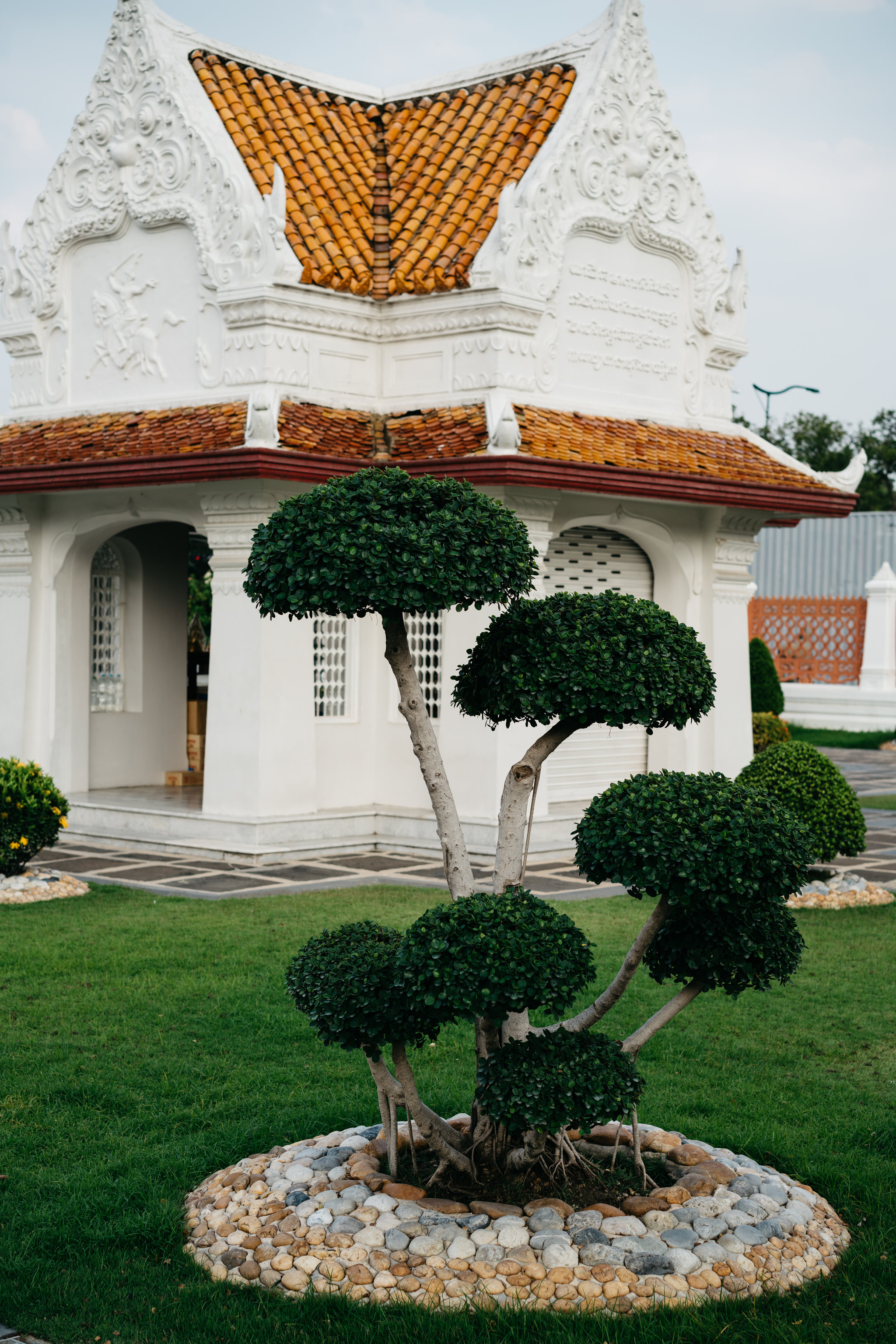 Ornate white pavilion with orange roof, and manicured tree in front.