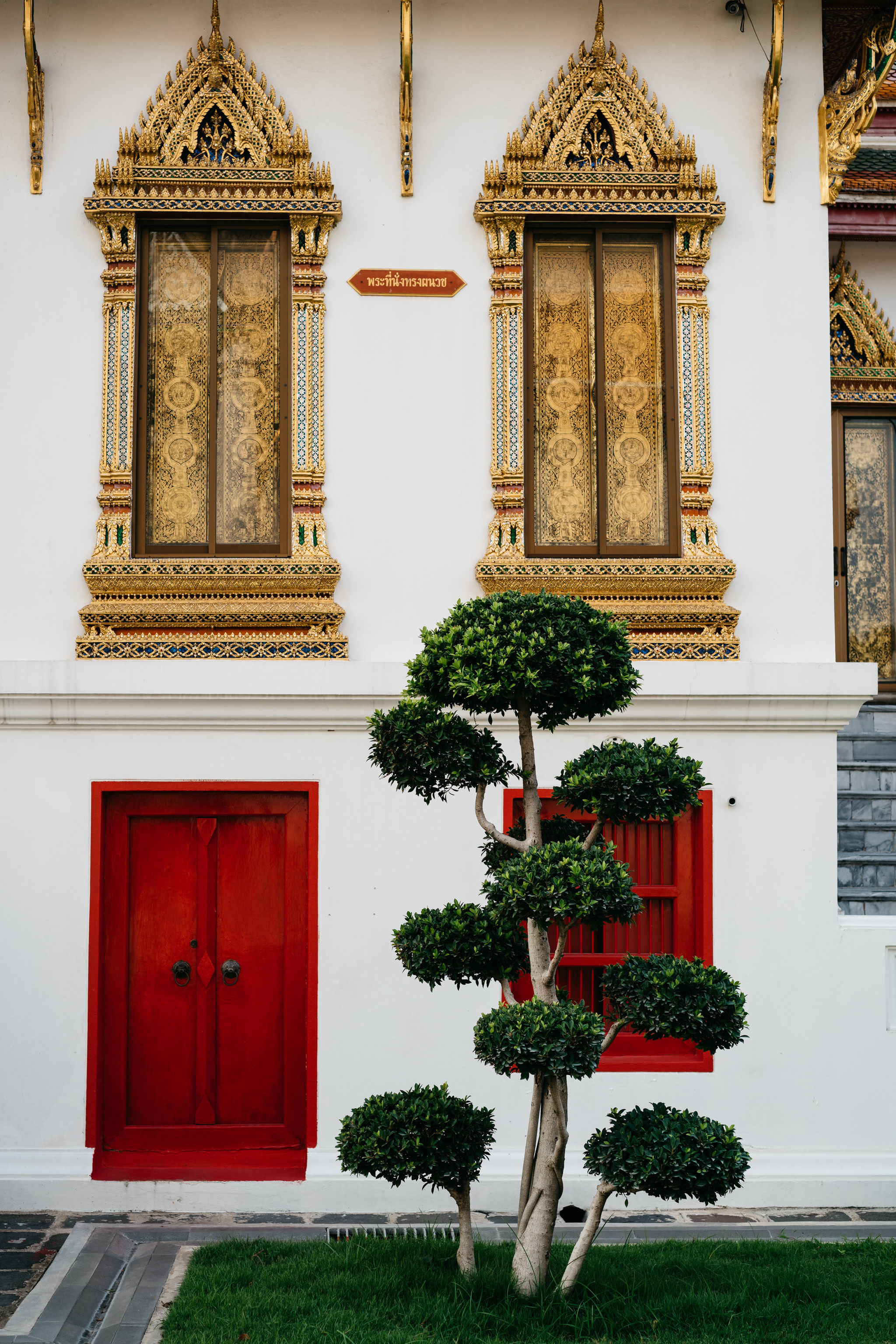 Wat Benchamabophit temple exterior, featuring ornate gold windows, a red door, and manicured shrubs.