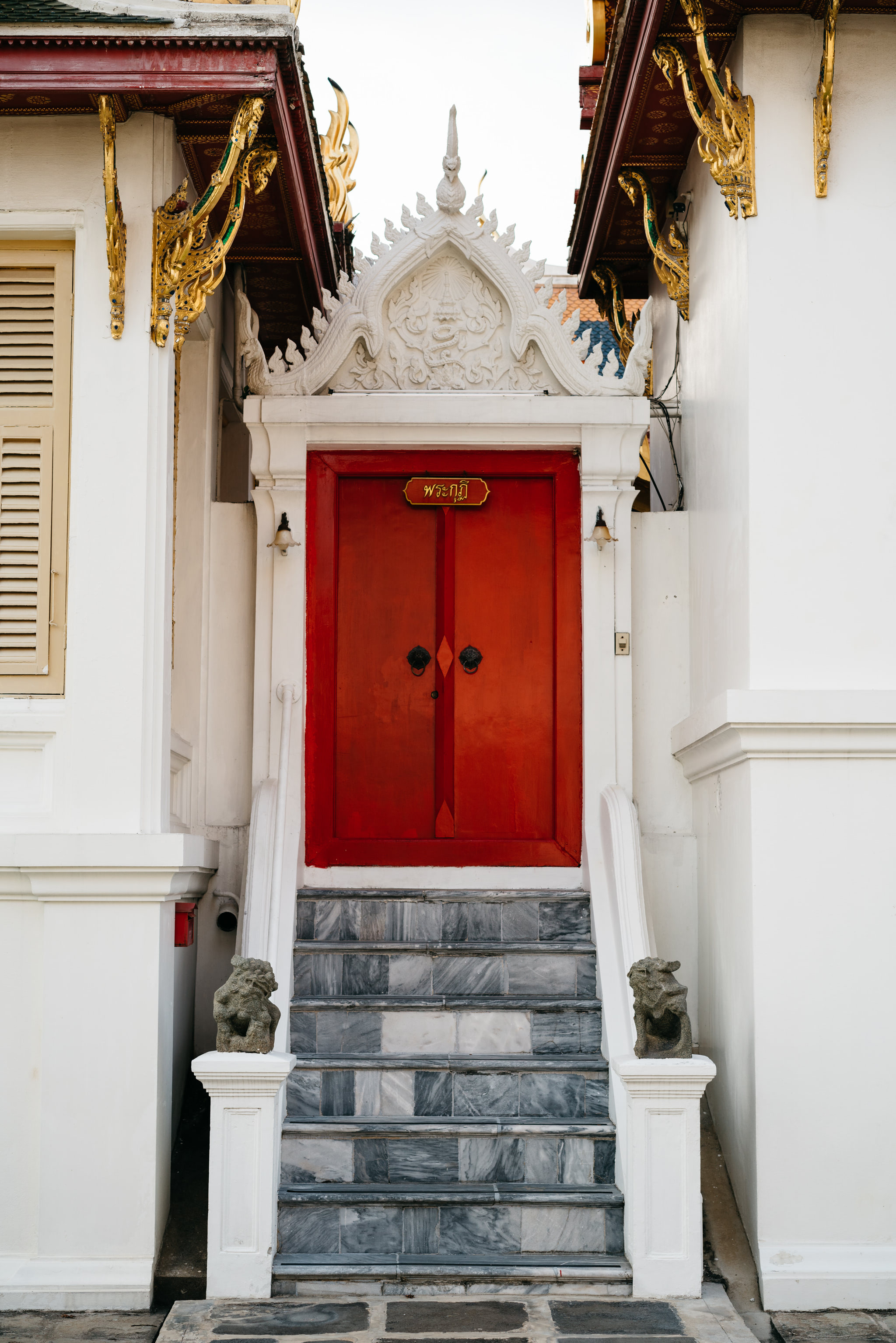 Red door with Thai script, marble steps, and ornate white and gold temple architecture.