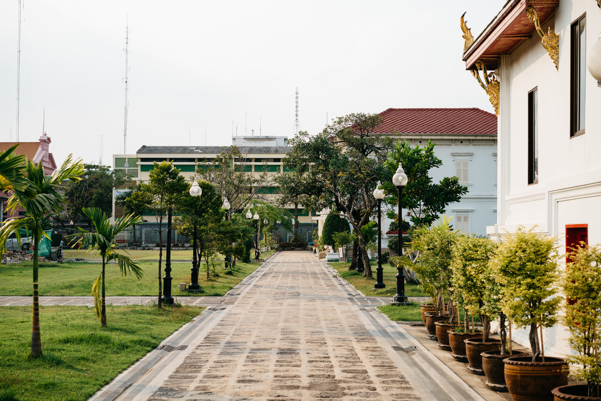 Stone pathway lined with potted plants and lamp posts leading to buildings in Bangkok.