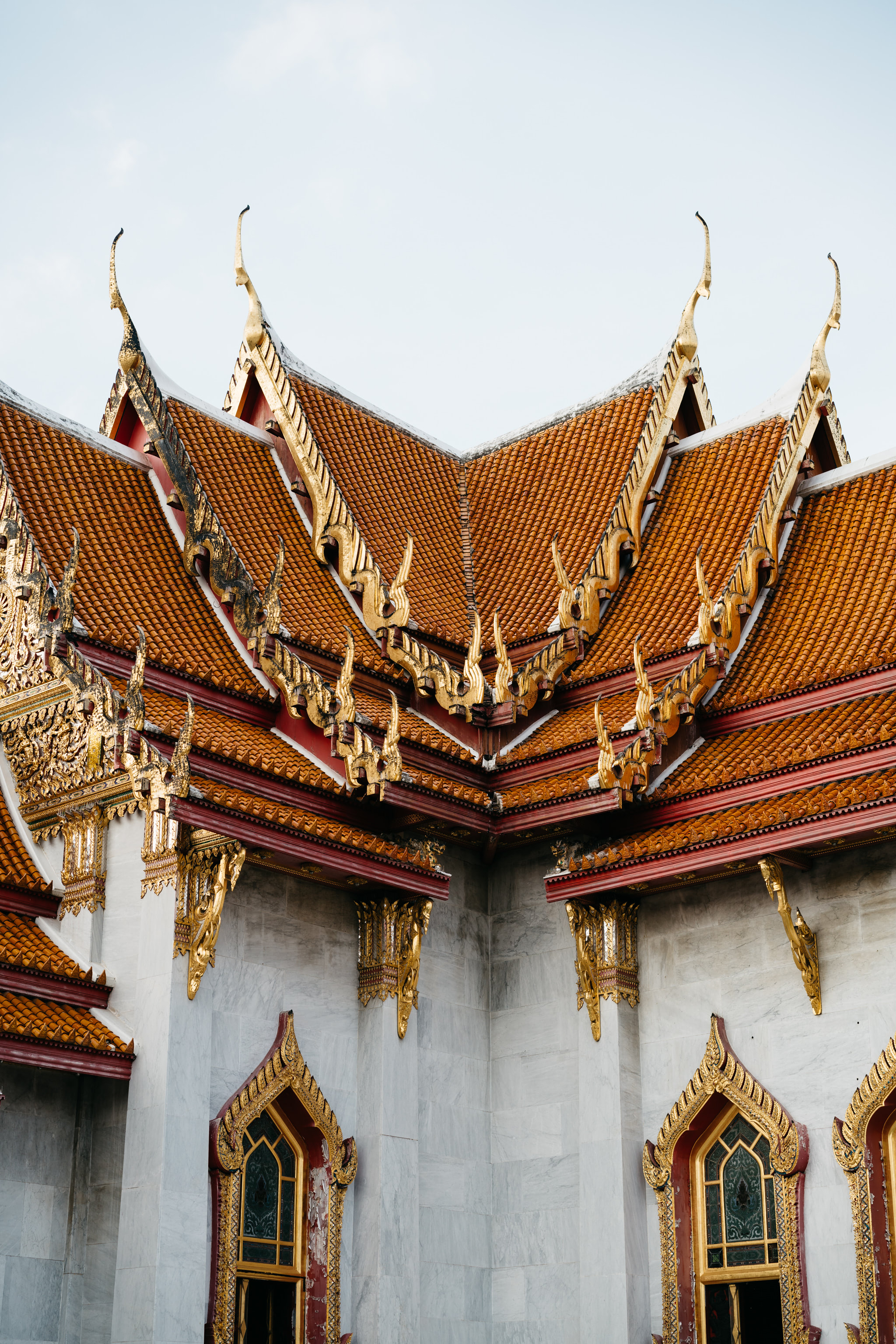 Ornate corner of Wat Benchamabophit temple in Bangkok, featuring a red and orange tiled roof with gold detailing and marble walls with gold-trimmed windows.