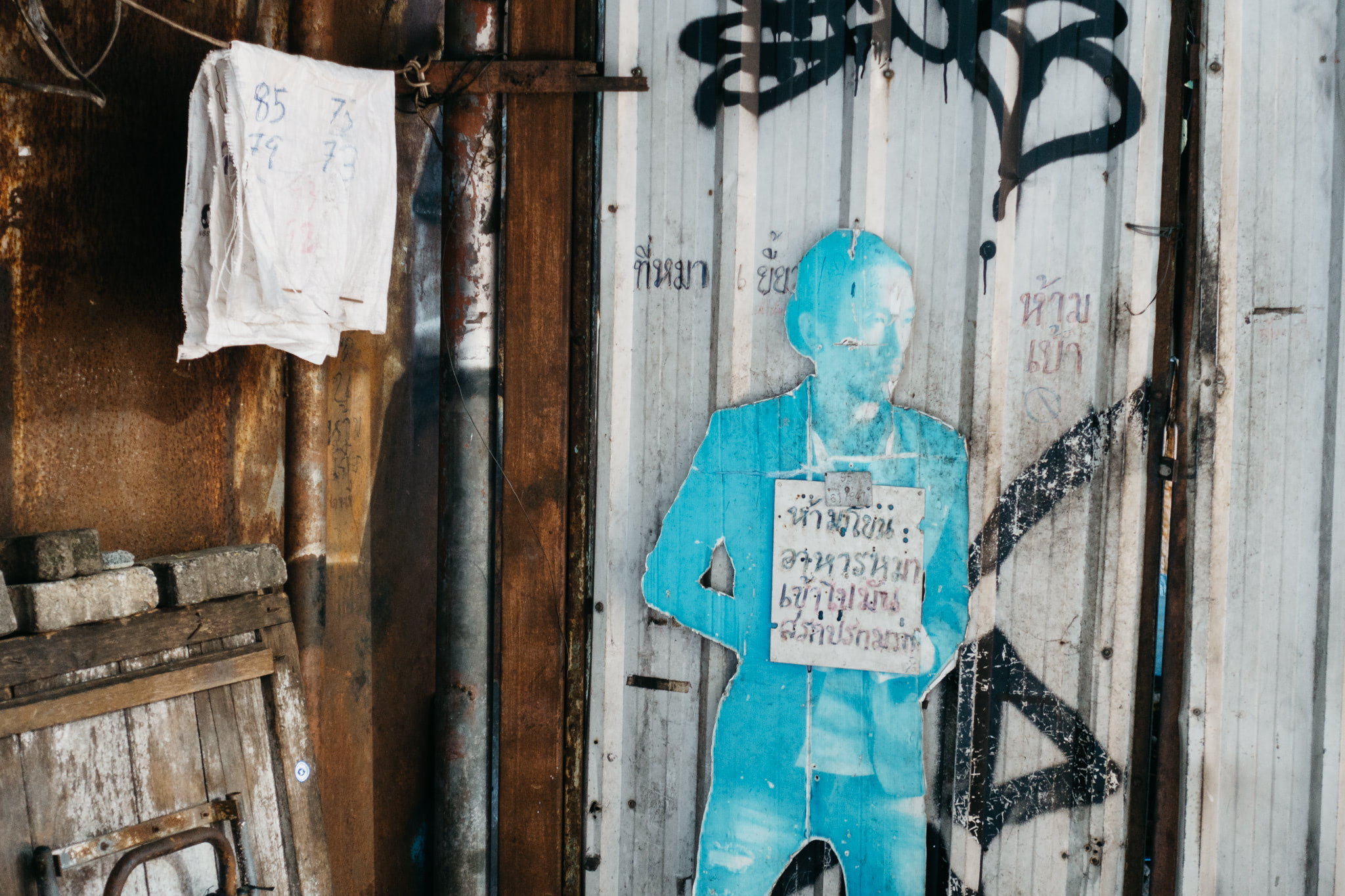 A faded blue cardboard cutout of a person stands against a rusty corrugated metal wall with graffiti and handwritten Thai text.
