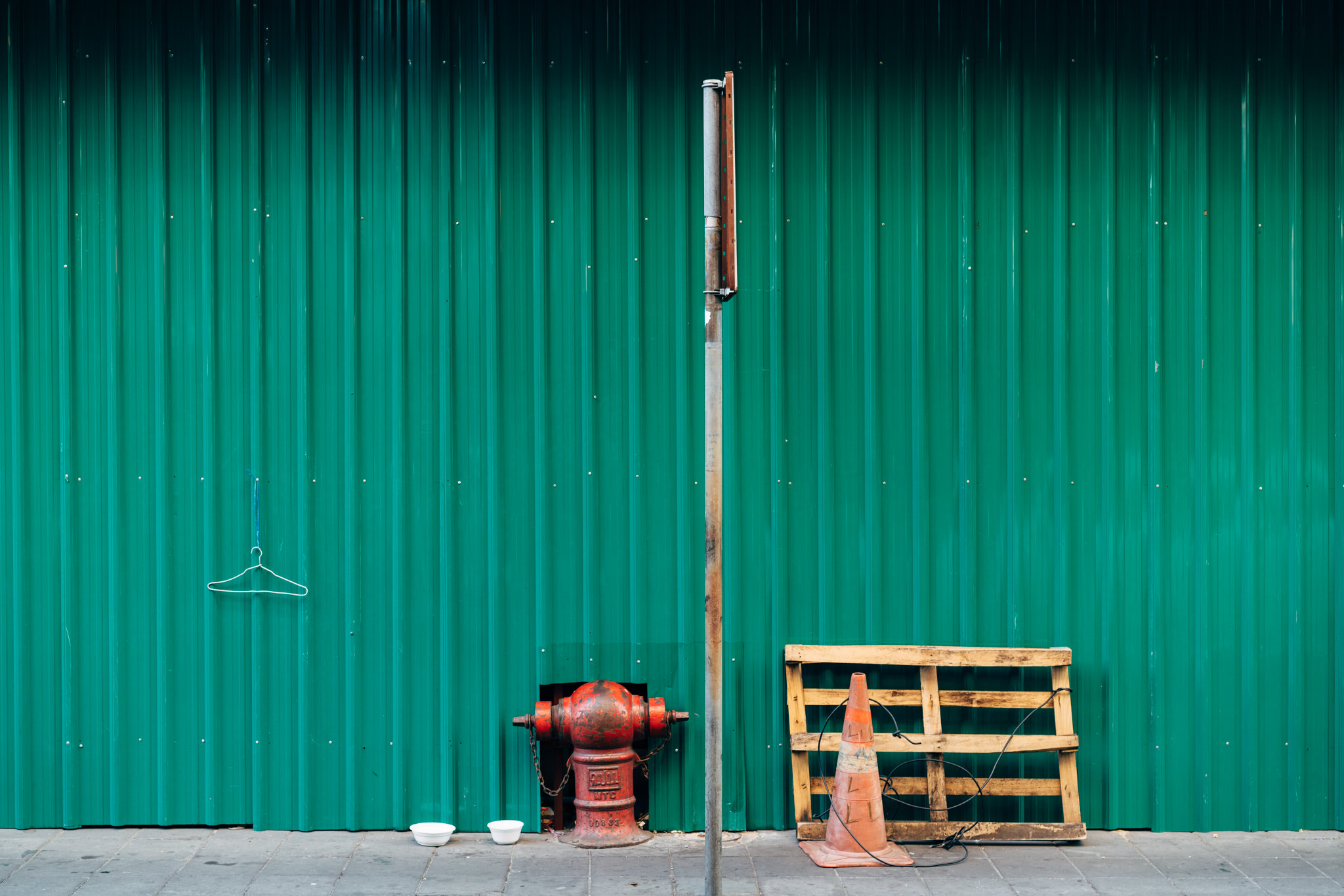 Green corrugated metal wall with fire hydrant, pallet, traffic cone, and clothes hanger.