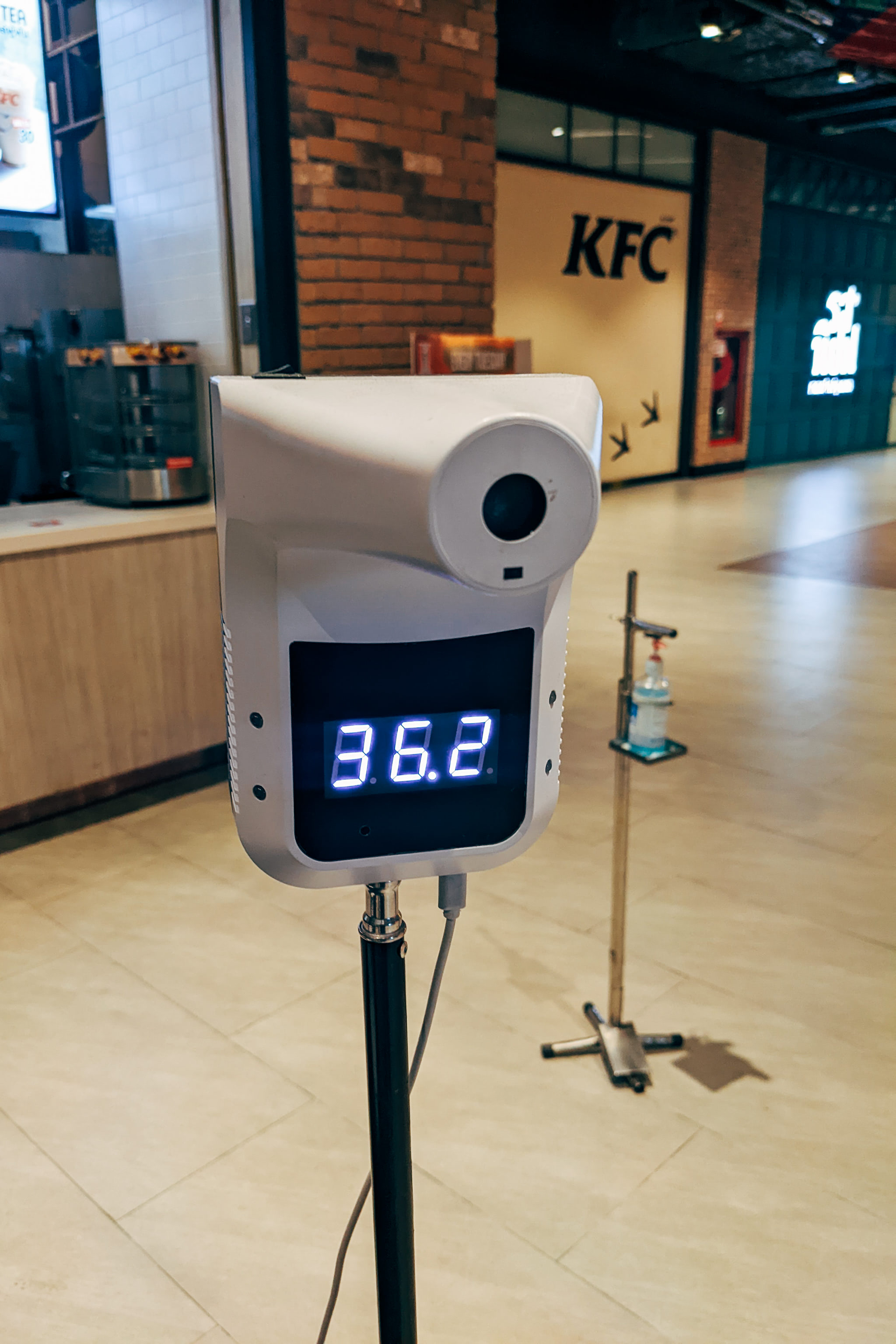 Thermal scanner displaying 36.2, mounted on a stand with a hand sanitizer dispenser nearby.