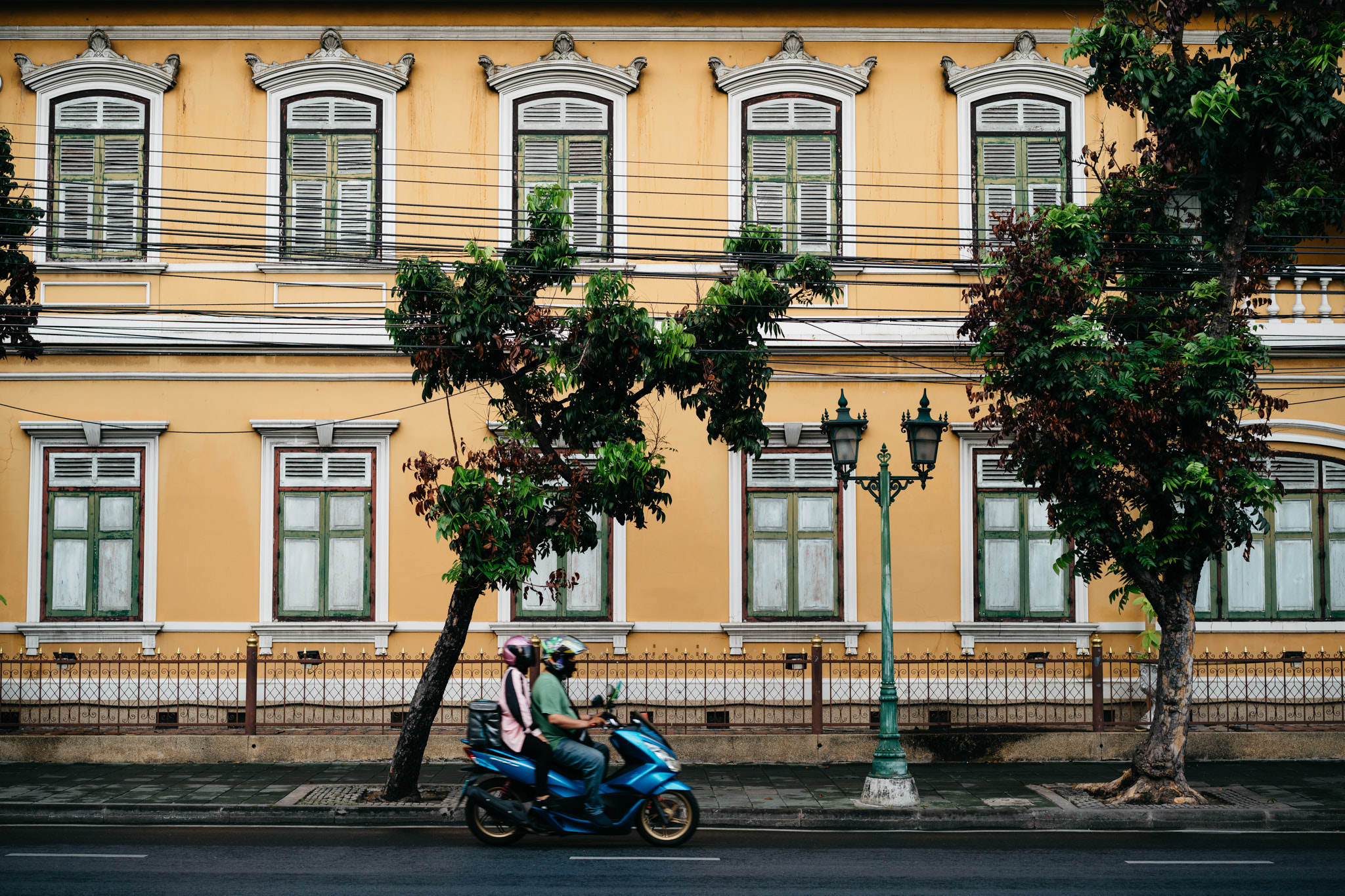 Two people on a blue scooter pass a yellow building with ornate windows and green trees.