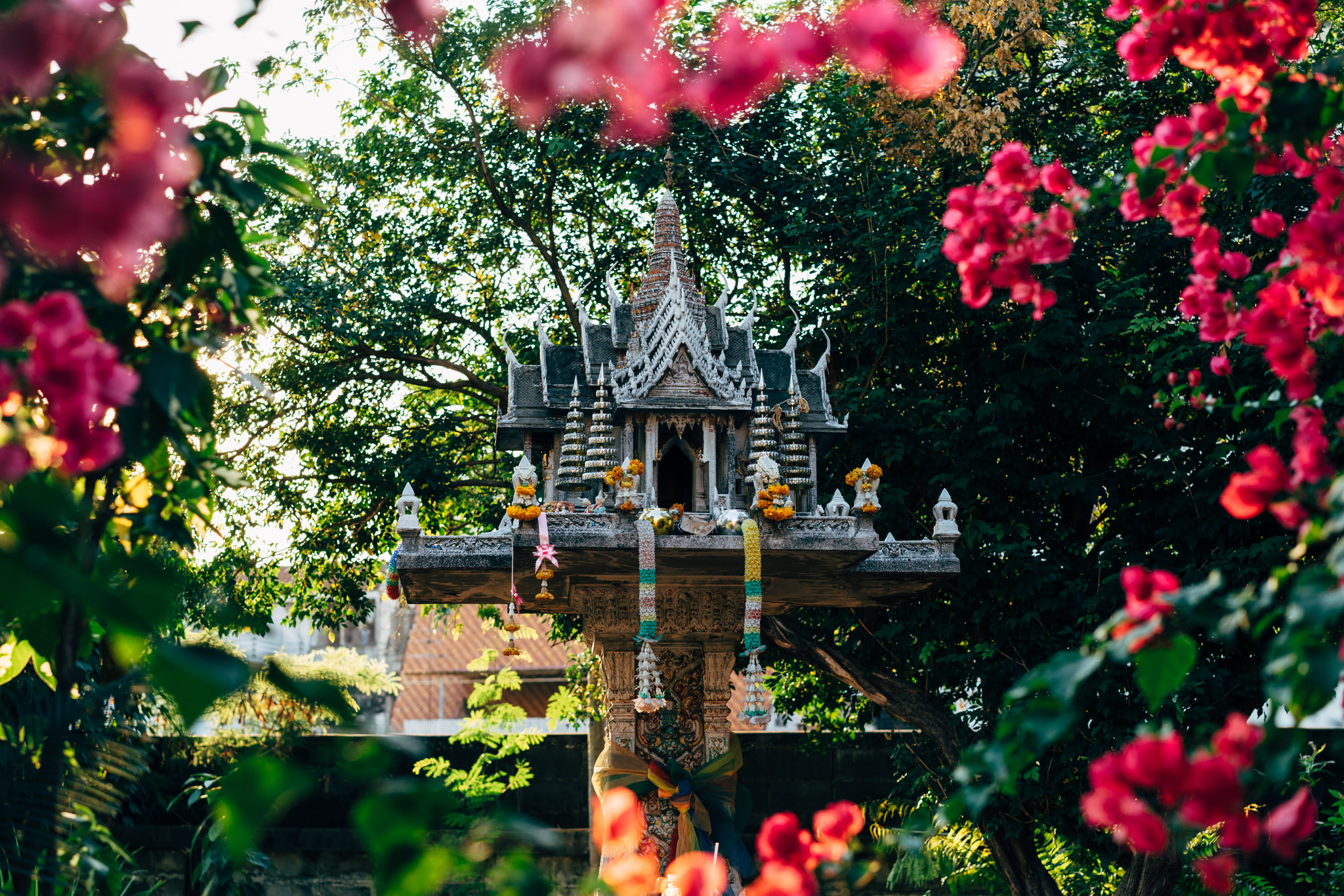 Ornate Thai shrine adorned with flowers, partially obscured by foliage.