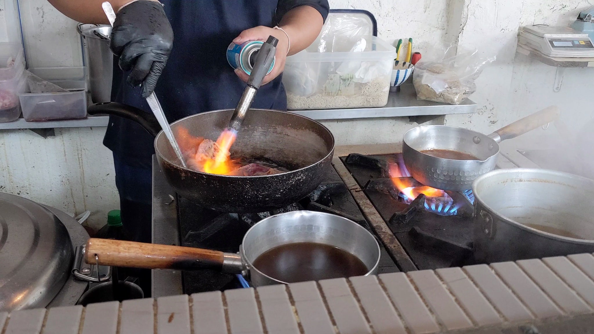 Chef torching meat in a pan on a gas stove.