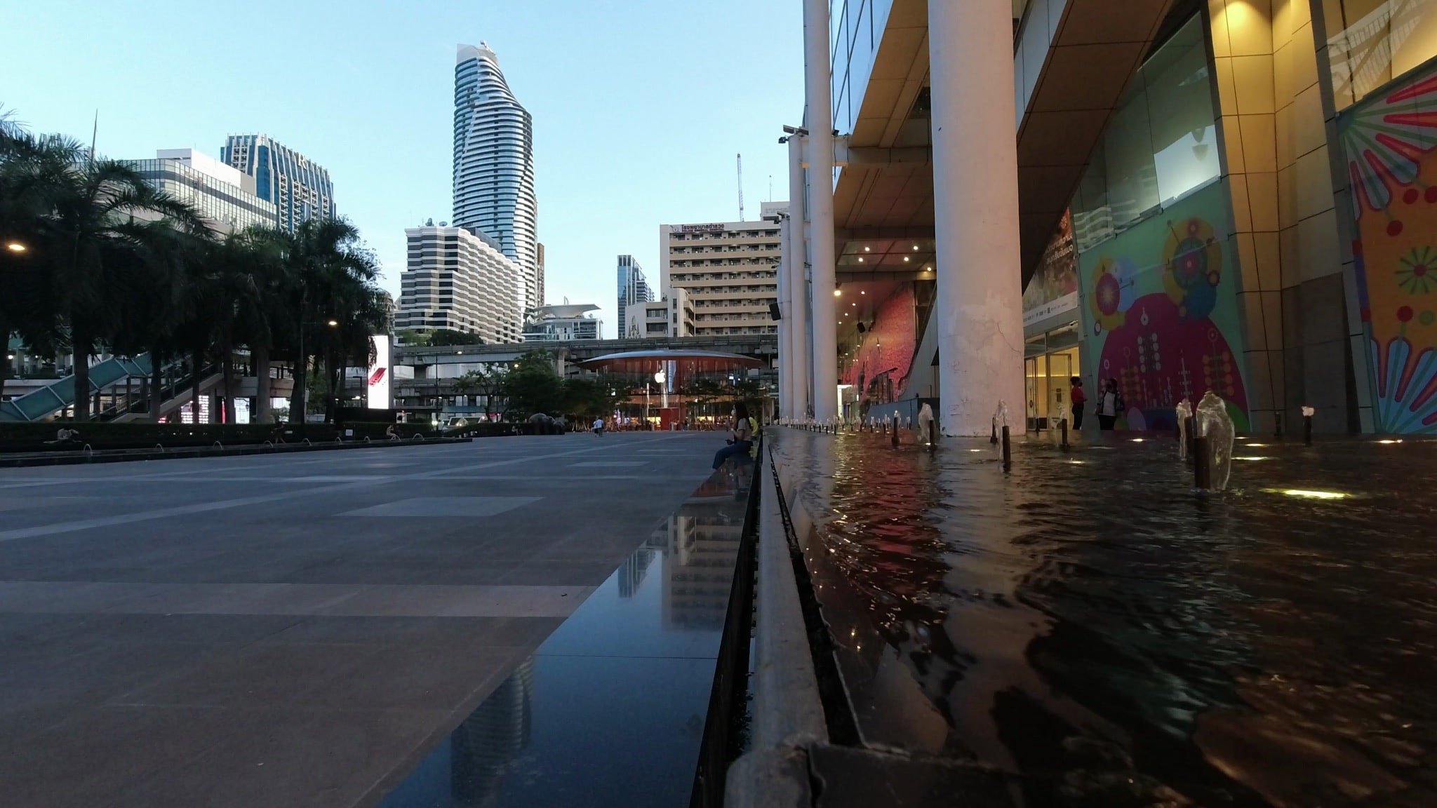 Central Bangkok fountain at dusk, reflecting city buildings.