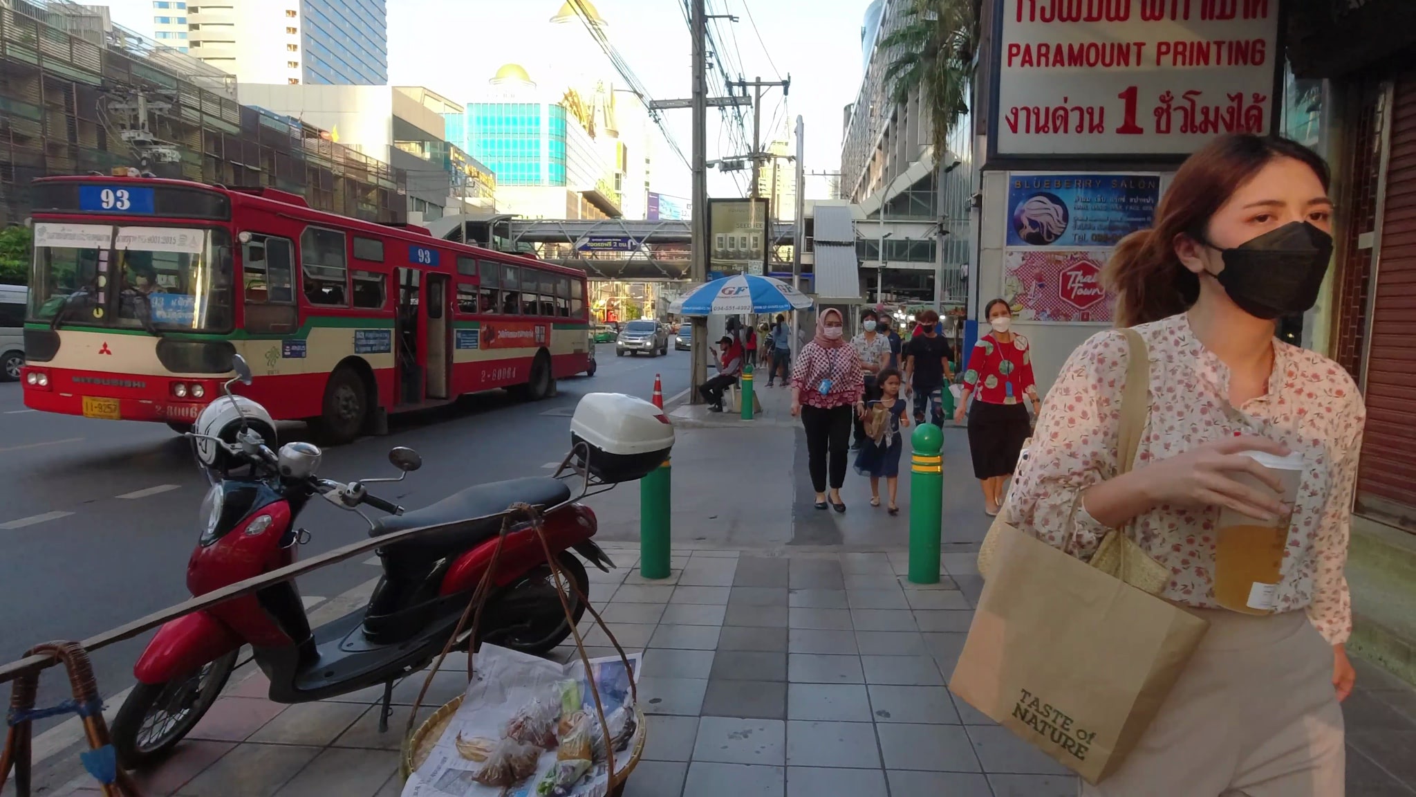 Woman in facemask walks Bangkok sidewalk past bus and parked scooter.