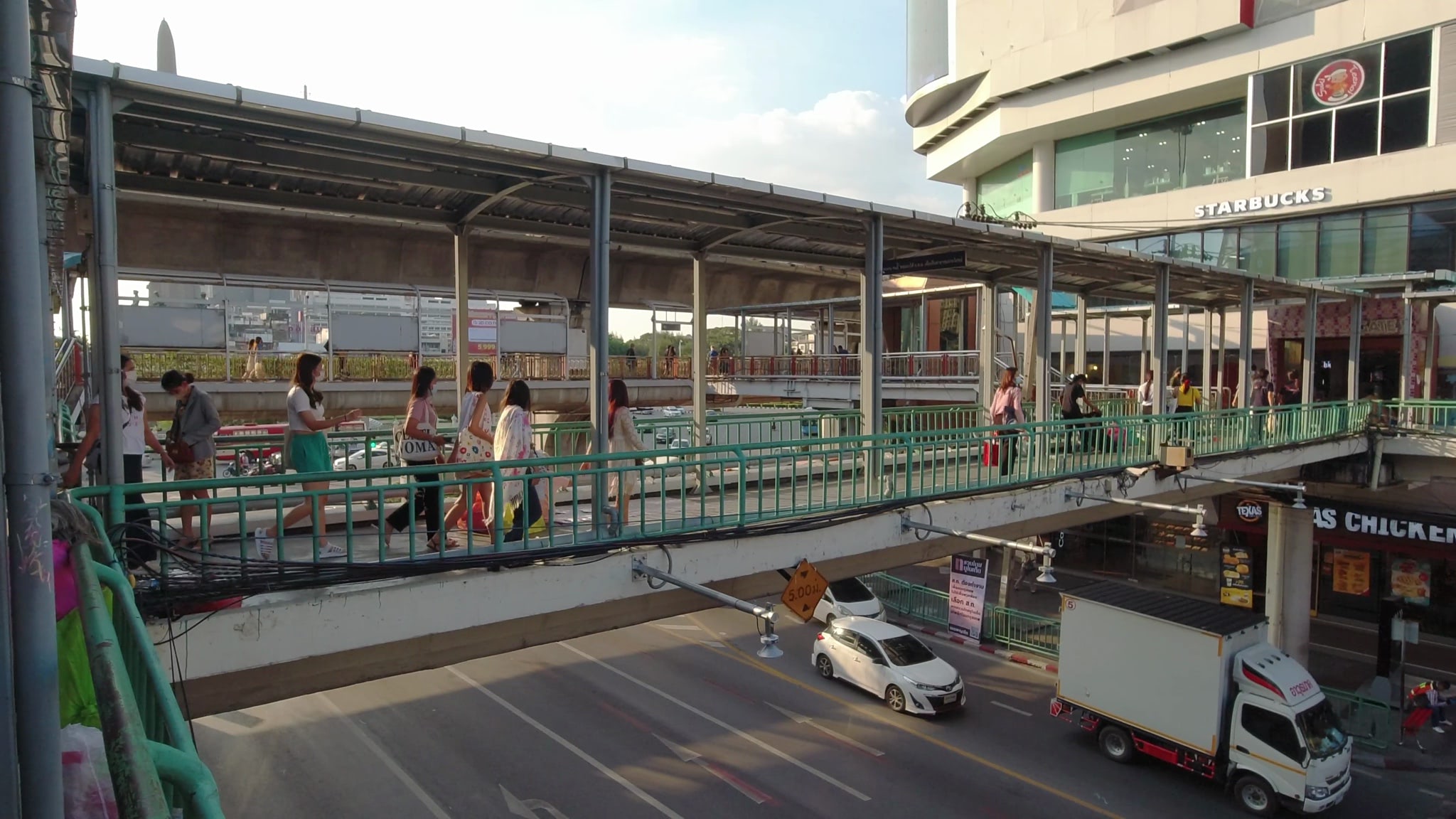 Pedestrians walking on a pedestrian bridge over a busy Bangkok street.