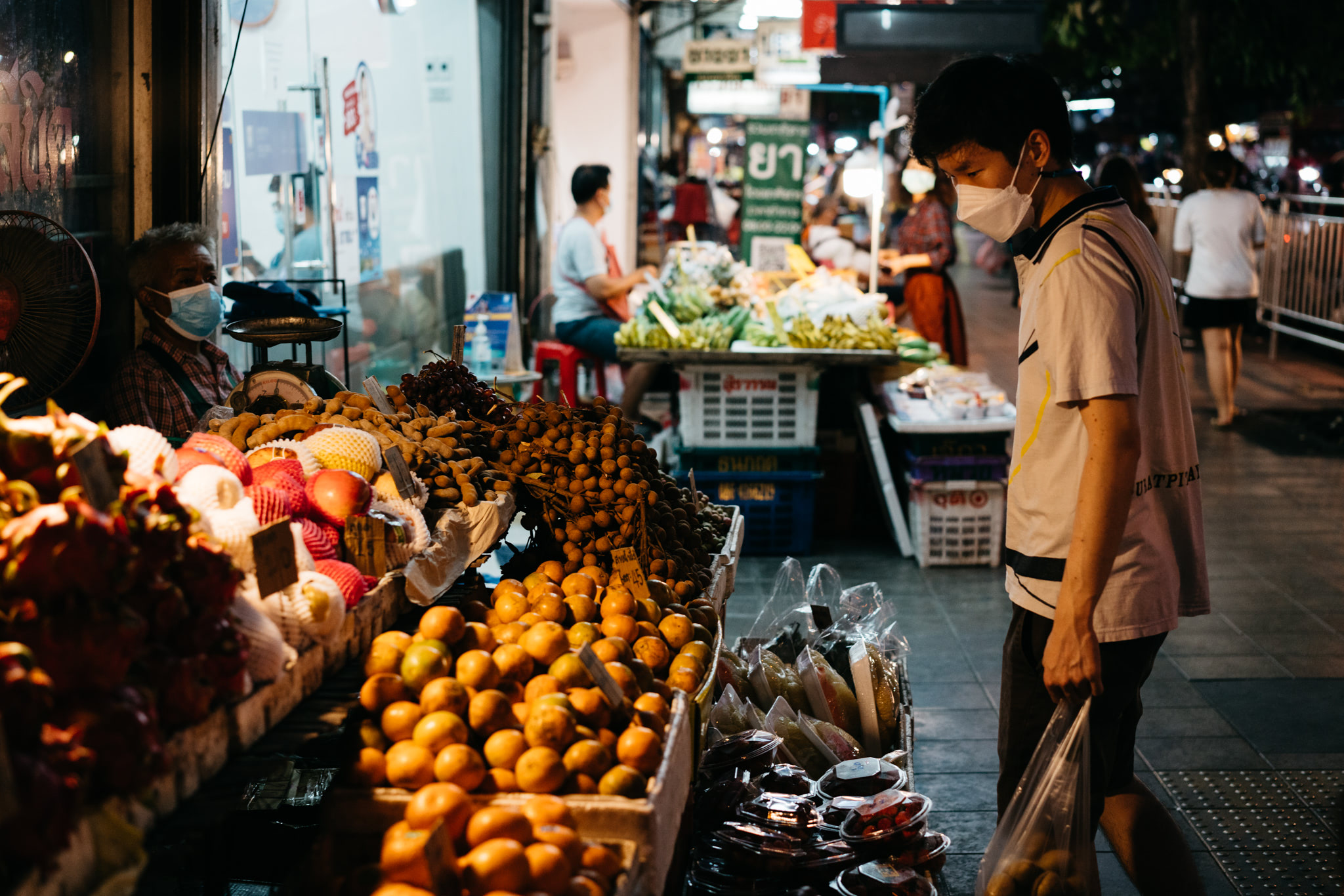 A man wearing a face mask shops at a Bangkok fruit stand overflowing with dragon fruit, mangosteen, oranges, and other tropical fruits.