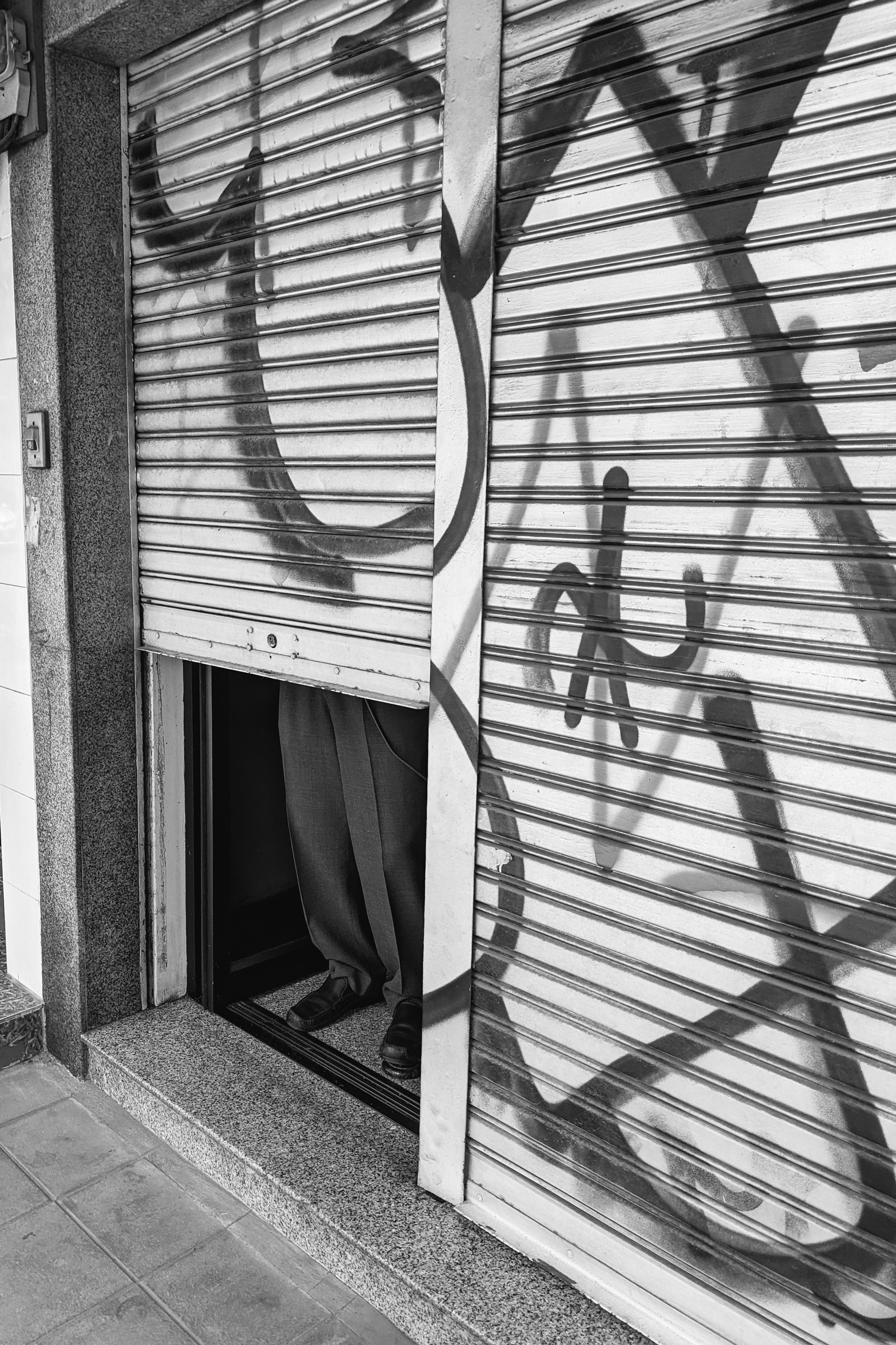 Black and white photo of a person's legs and feet visible beneath a partially raised metal security gate with graffiti.