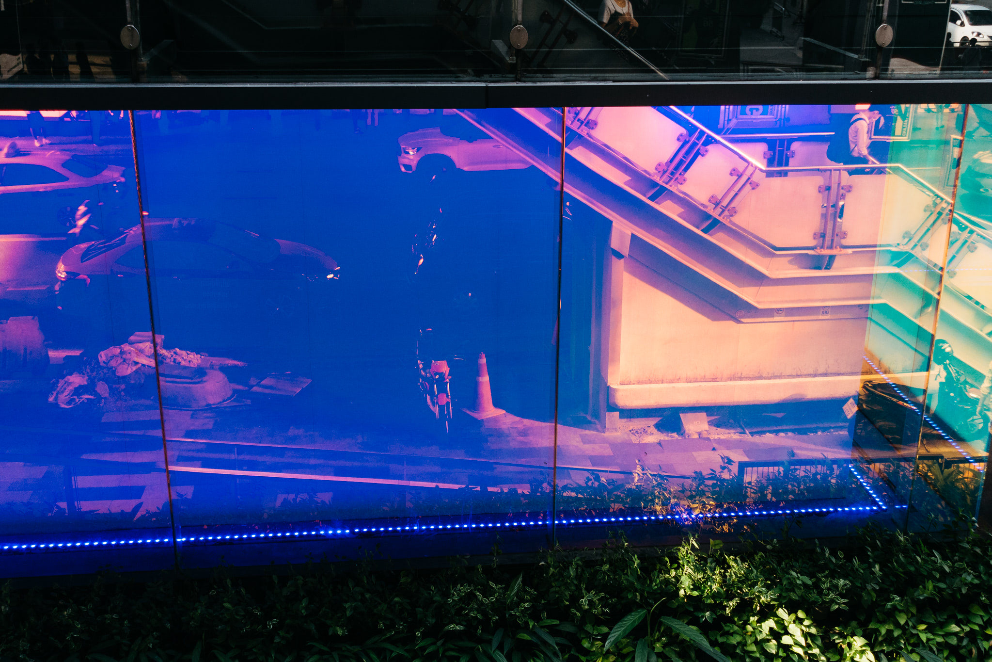Abstract image of a building exterior at night, showing distorted reflections of cars and a staircase in purple and blue tones, illuminated by blue LED lights at the base.