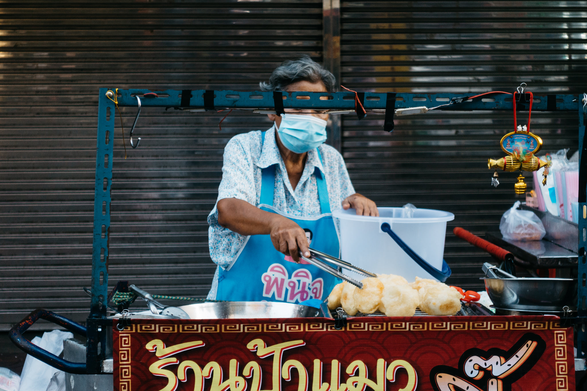 Bangkok street vendor wearing a mask, using tongs to cook food at a stand.