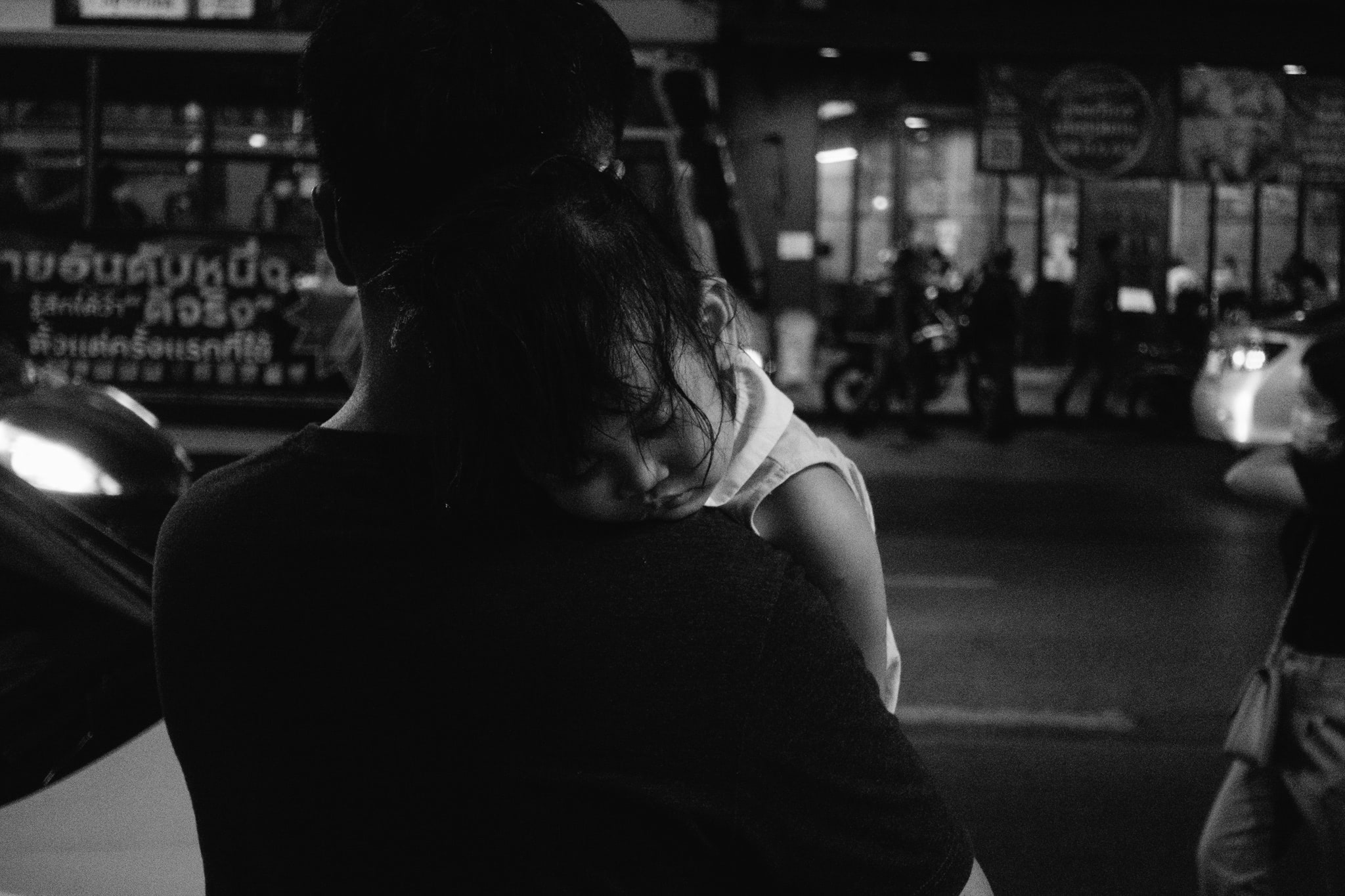 Black and white photo of a child sleeping on a person's shoulder in Chinatown at night.