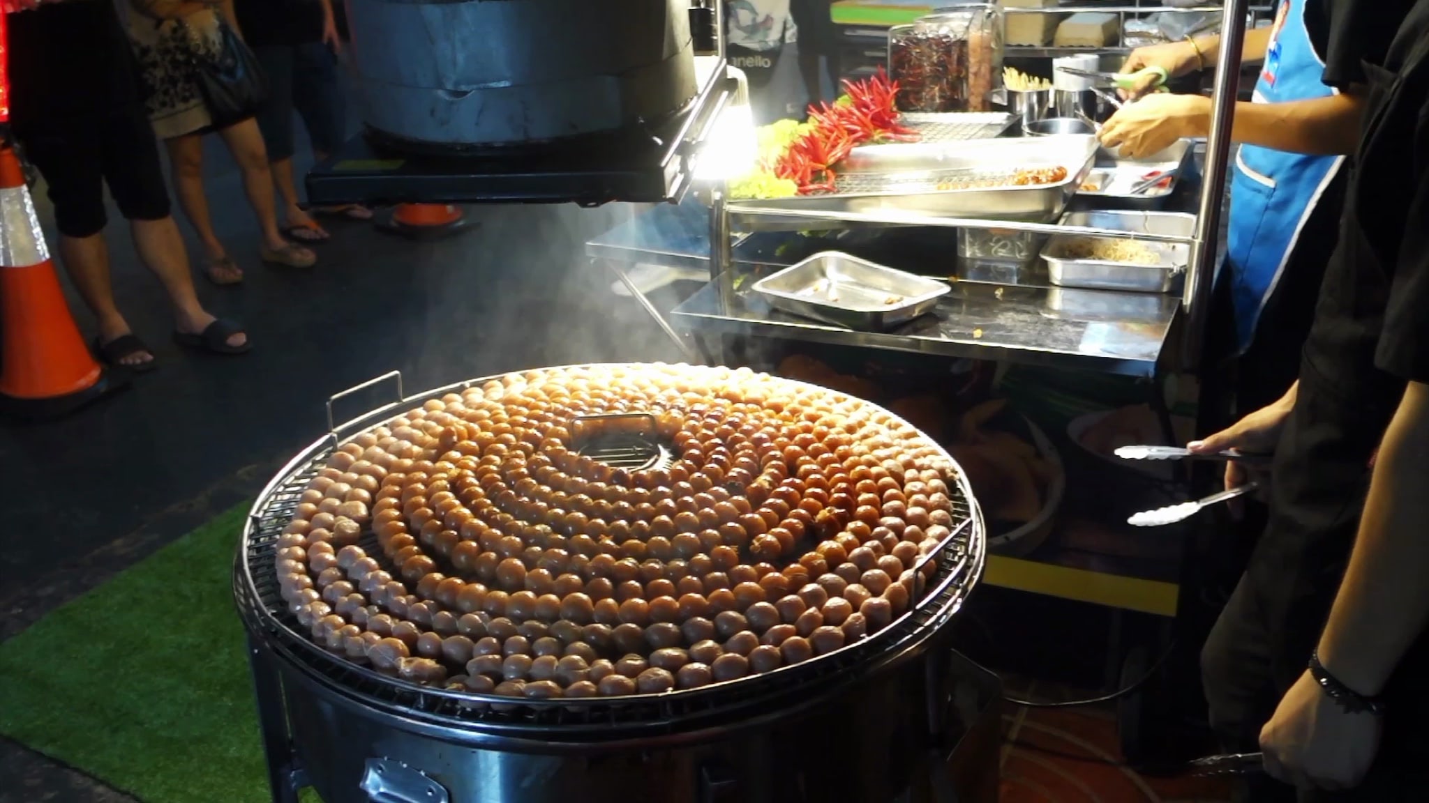 Many sausages grilling in a circular pattern on a large metal grill.