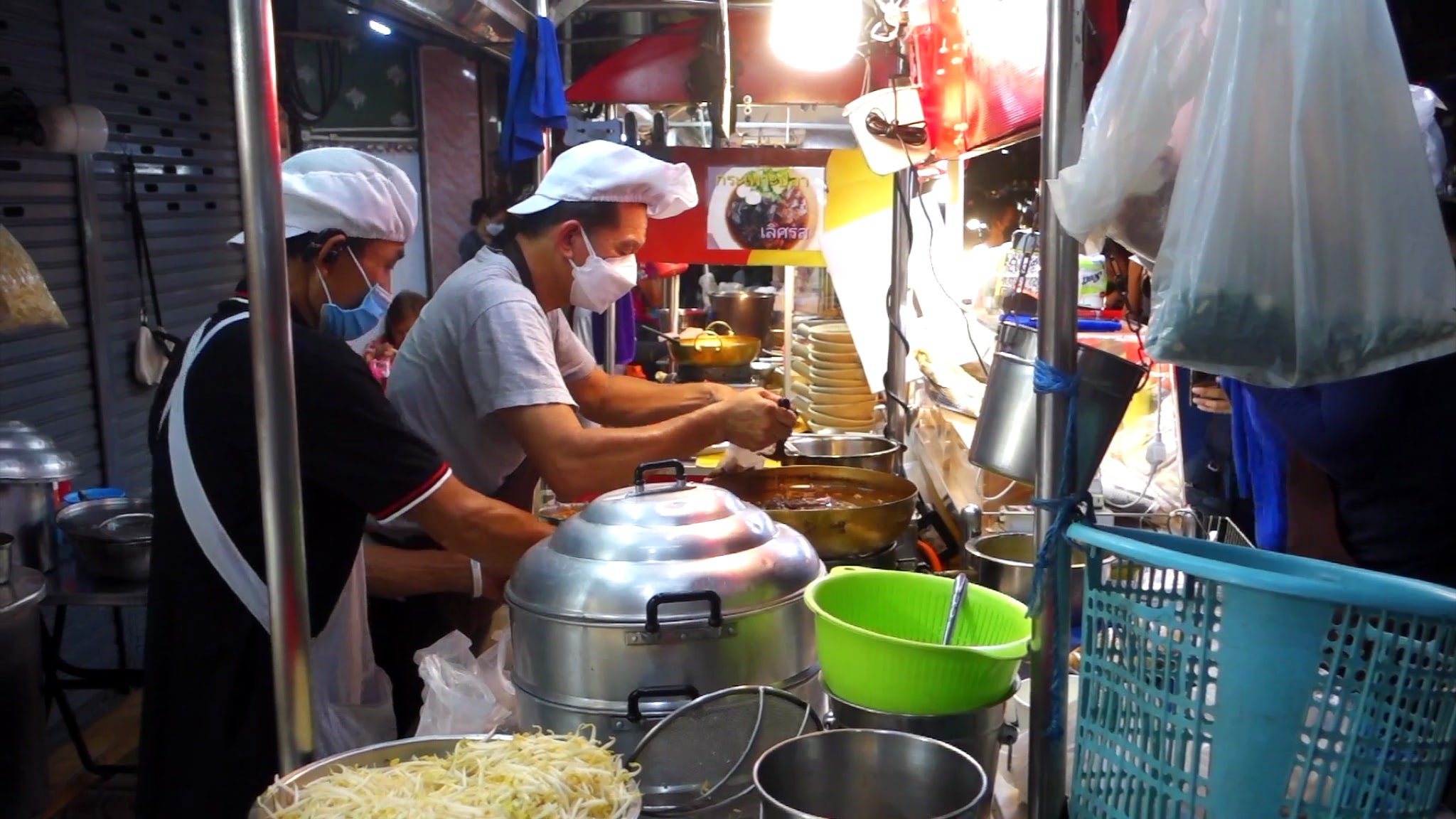 Two cooks wearing masks prepare food at an outdoor food stall.