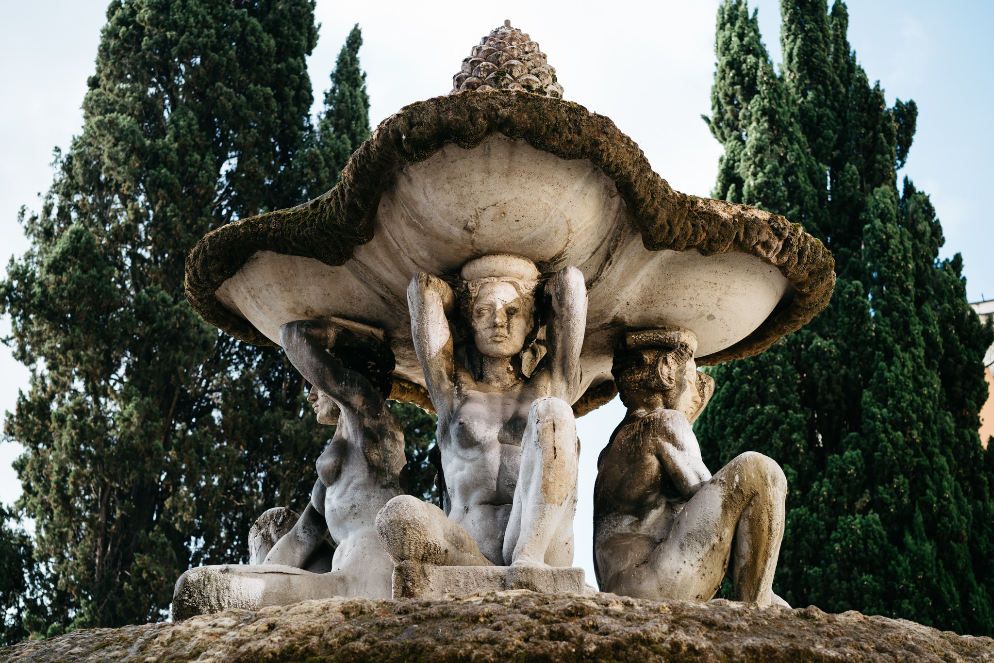 Stone fountain in Rome featuring three female figures supporting a large, moss-covered basin.