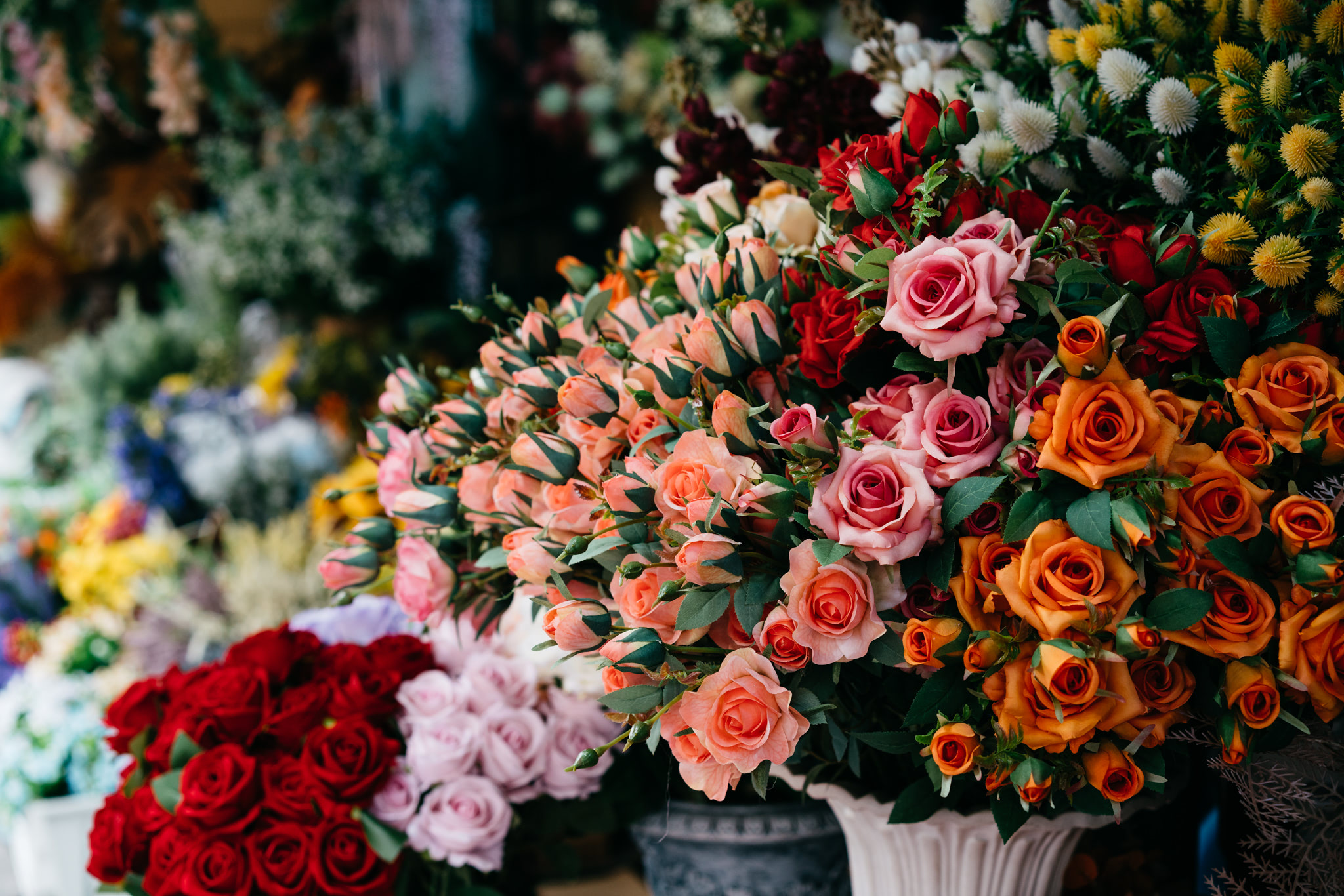 Close-up of a bouquet of artificial roses in various colors, including pink, orange, and red, displayed in a vase at a flower market.