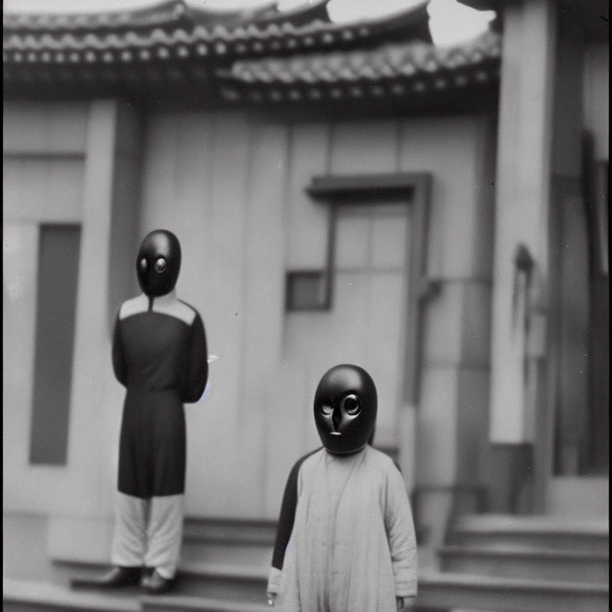 Black and white photo of two figures wearing black, oval-shaped masks in front of a building.