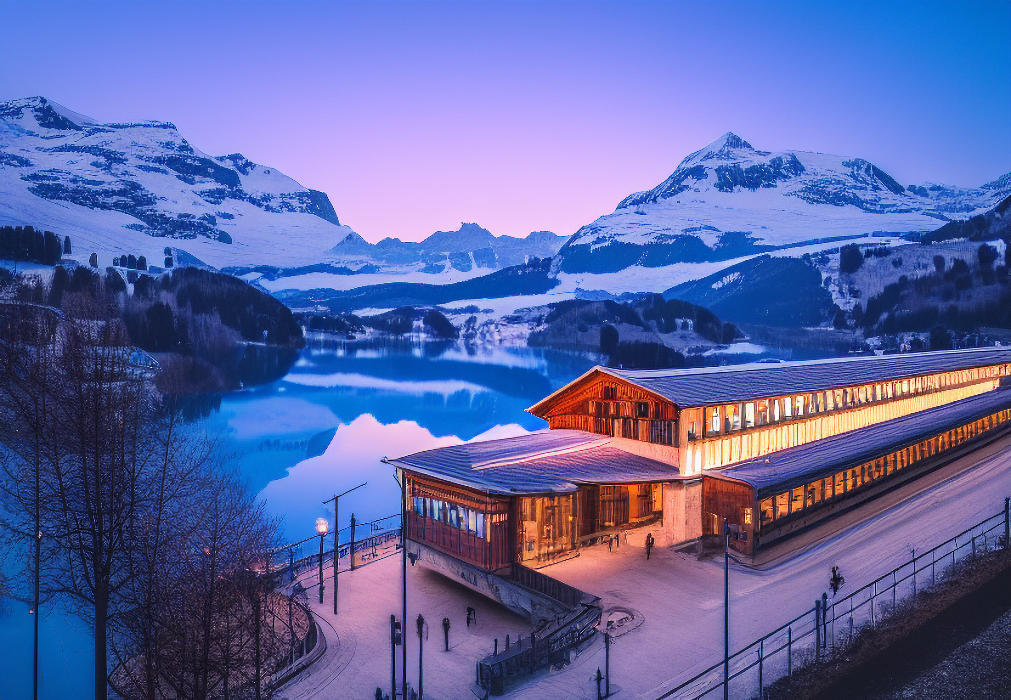 Lit-up Swiss train station at dusk, nestled in snowy Alps mountains beside a lake.