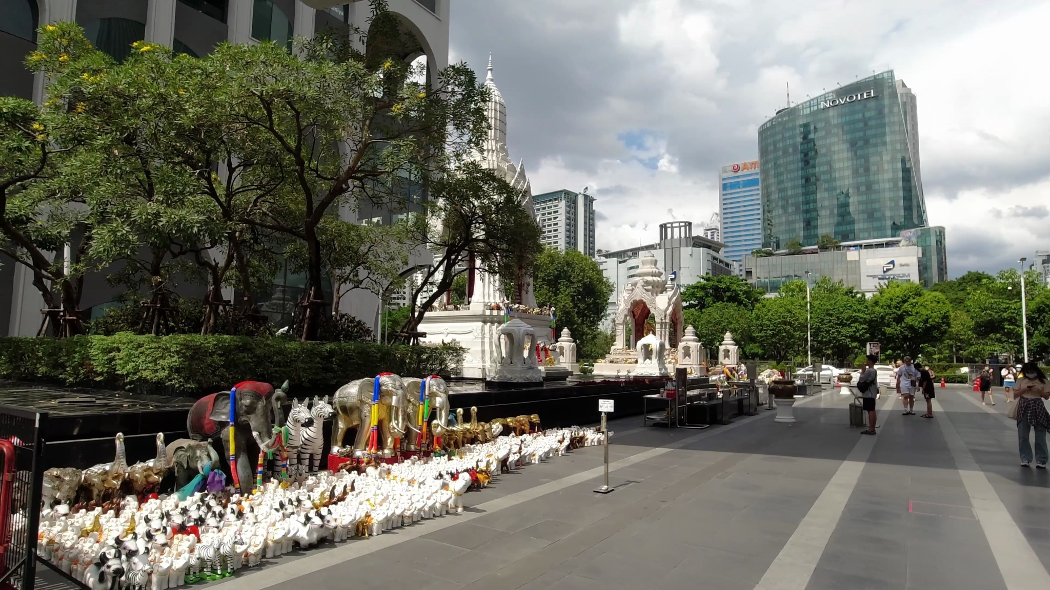 Bangkok shrine with elephant statues and offerings in front of a modern building.