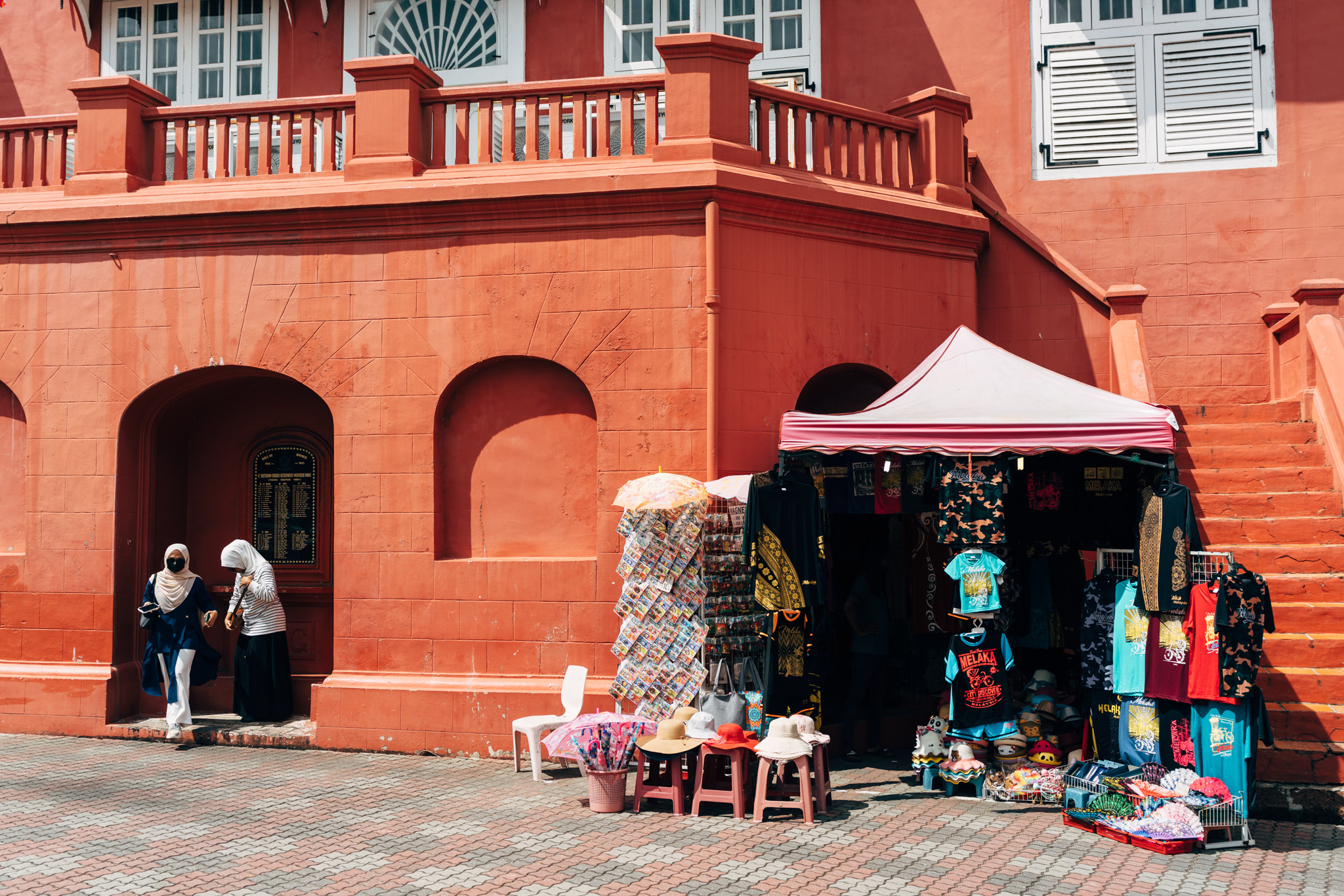Two women in hijabs walk past a souvenir stand in front of a red building in Malacca City.