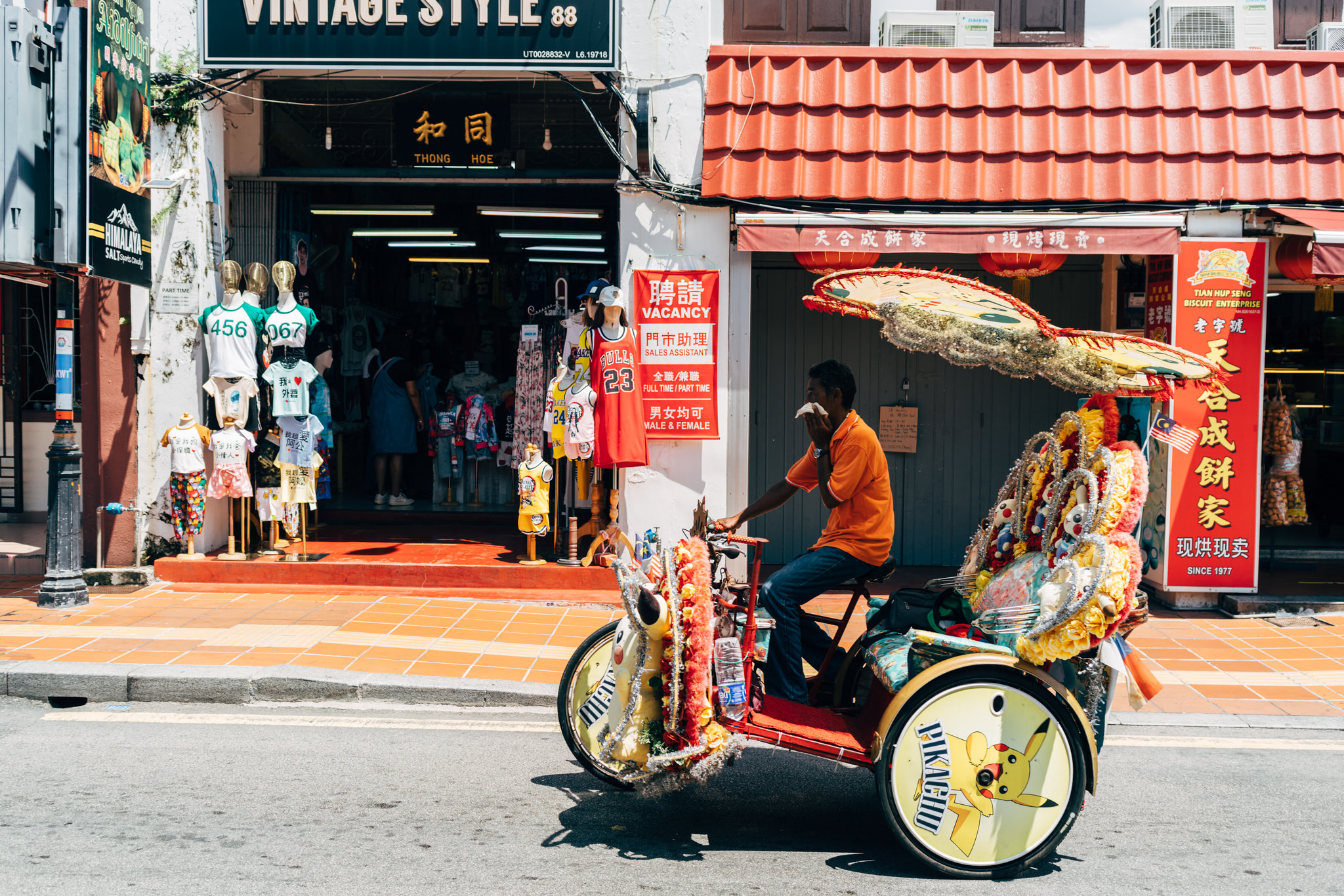 A man in an orange shirt driving a decorated trishaw with a Pikachu design on the wheel, parked on a street in Malacca, Malaysia.