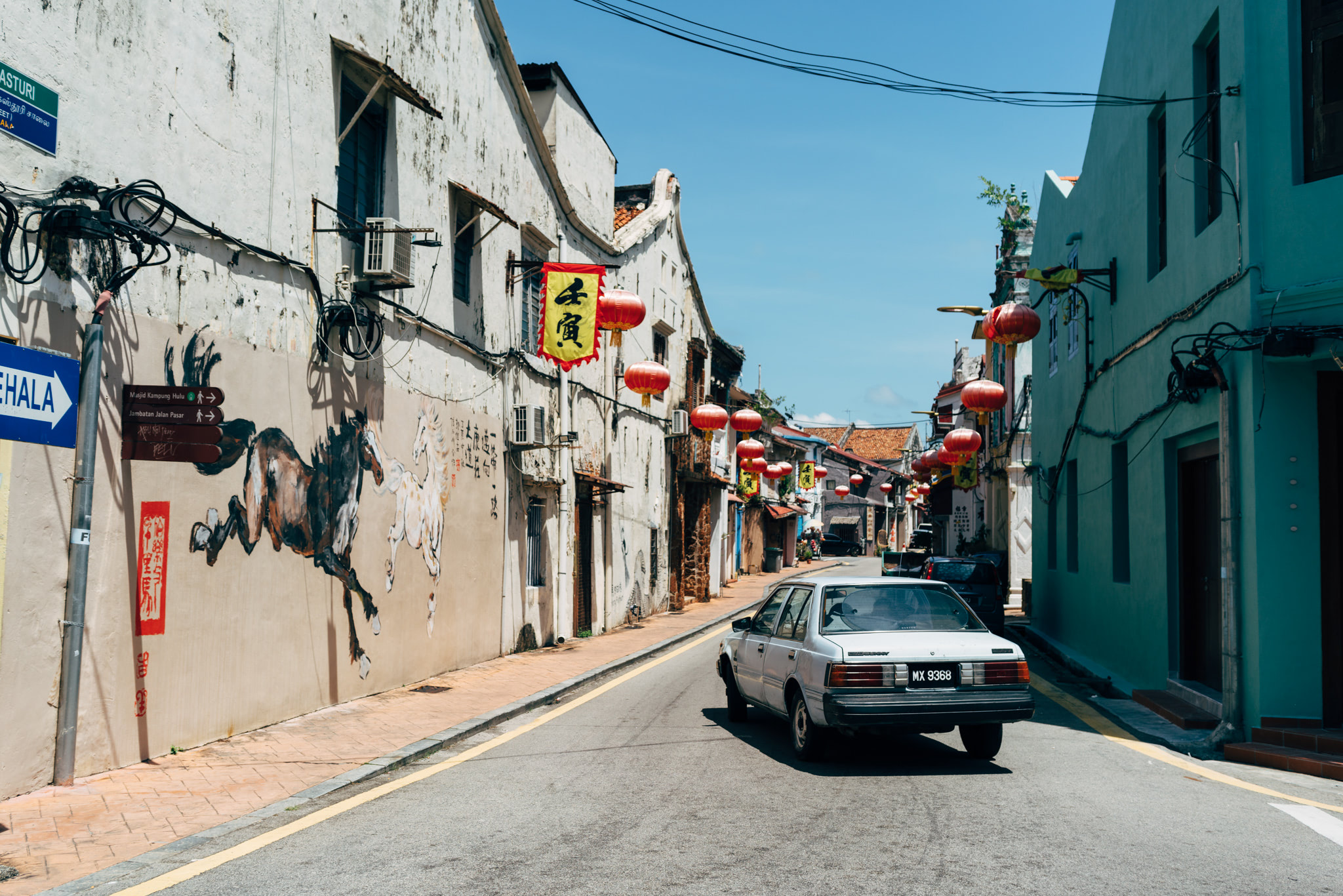 A silver car drives down a narrow street in Malacca, Malaysia, lined with aged buildings decorated with red lanterns and Chinese calligraphy.